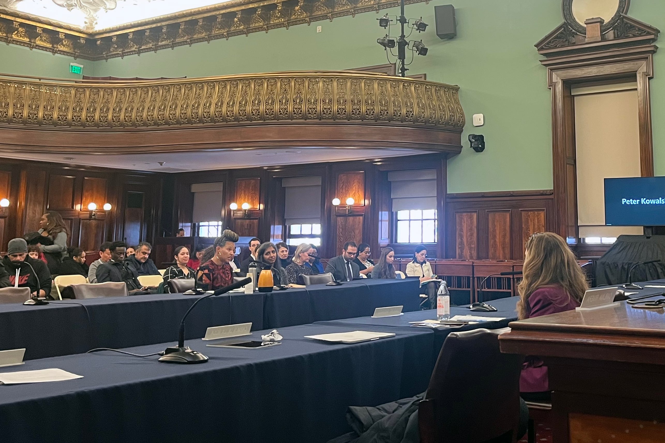 A group of people sit at a long wooden table in a conference room with people sitting in the foreground and background.