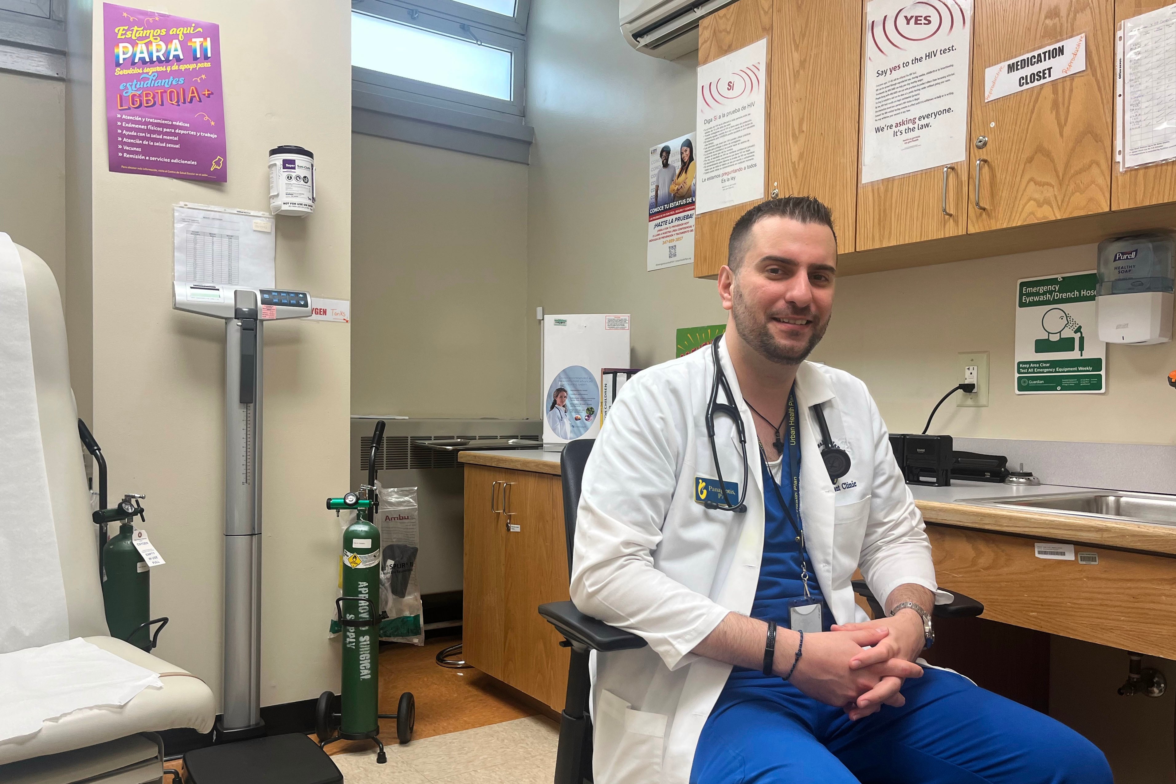 A man in a white dr's coat and blue scrubs sits in a chair in an exam room while smiling and posing for the camera.