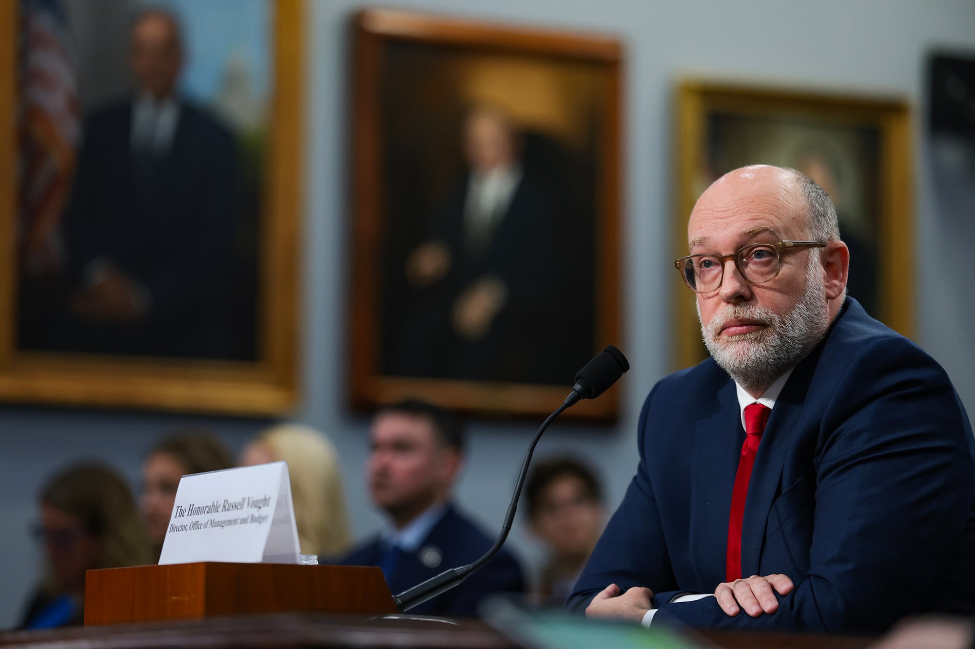 A white man in a suit sits at a table looking off from with a group of people sitting in chairs behind him during a House hearing on Capitol Hill.