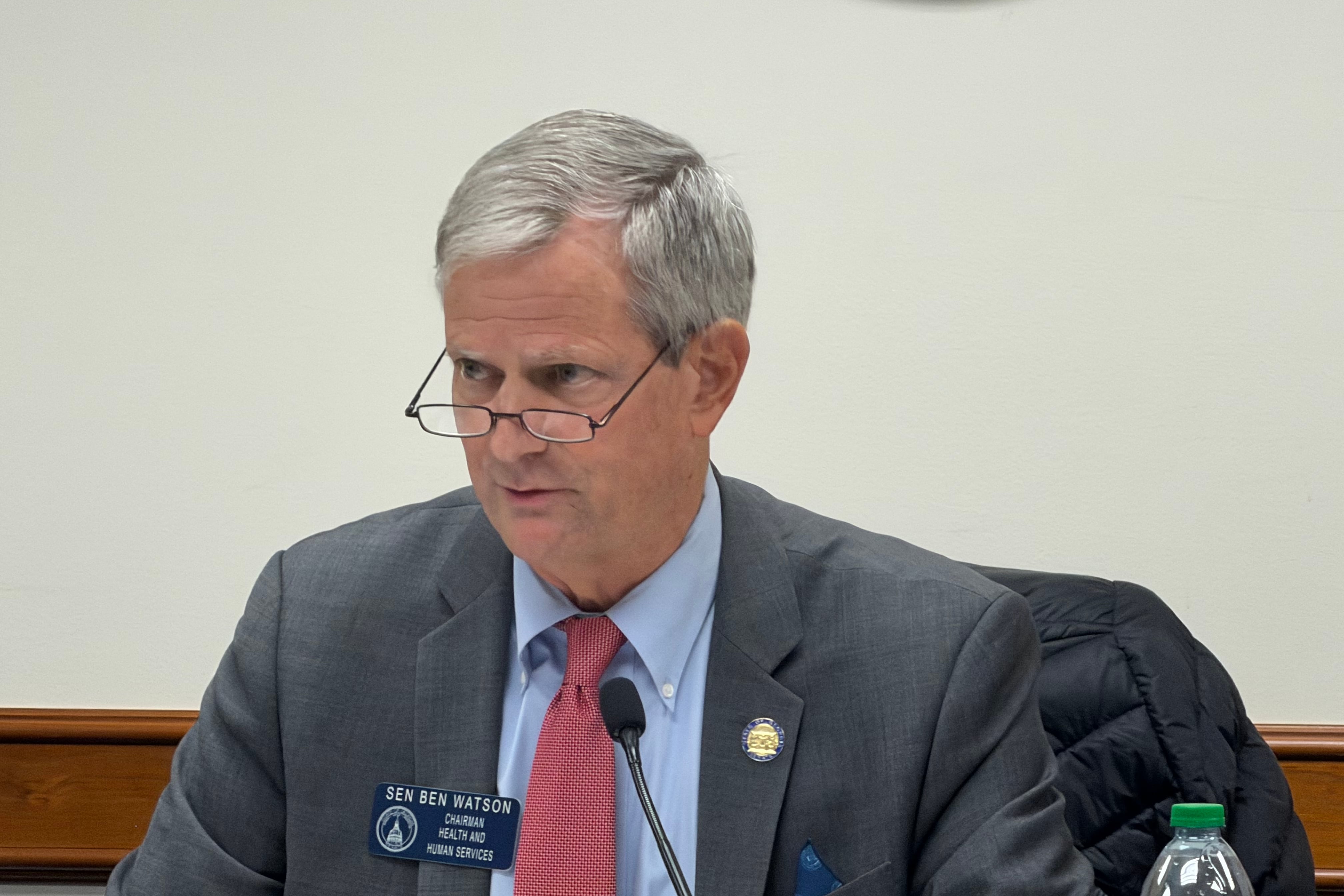 A man with gray hair and glasses wearing a gray suit and red tie sits behind a microphone.