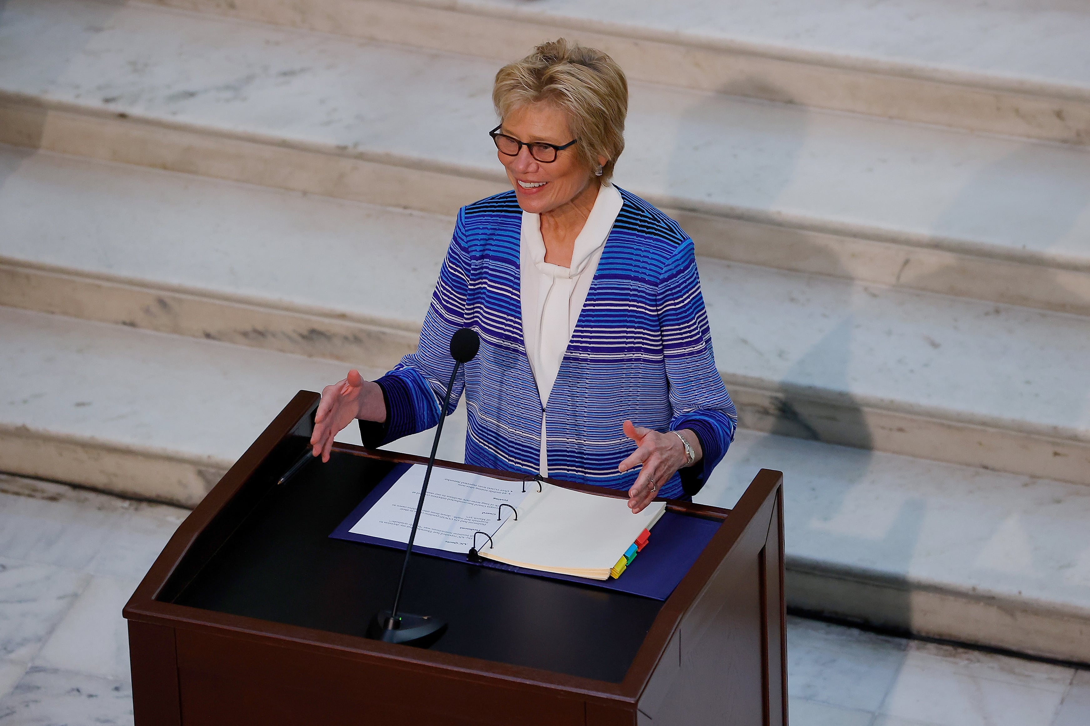 A white woman in a business suit speaks from behind a wooden podium and in front of a large set of stone stairs.