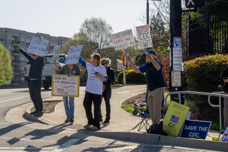 A small group of people stand on a sidewalk outside of a building on a sunny day holding signs during a protest.