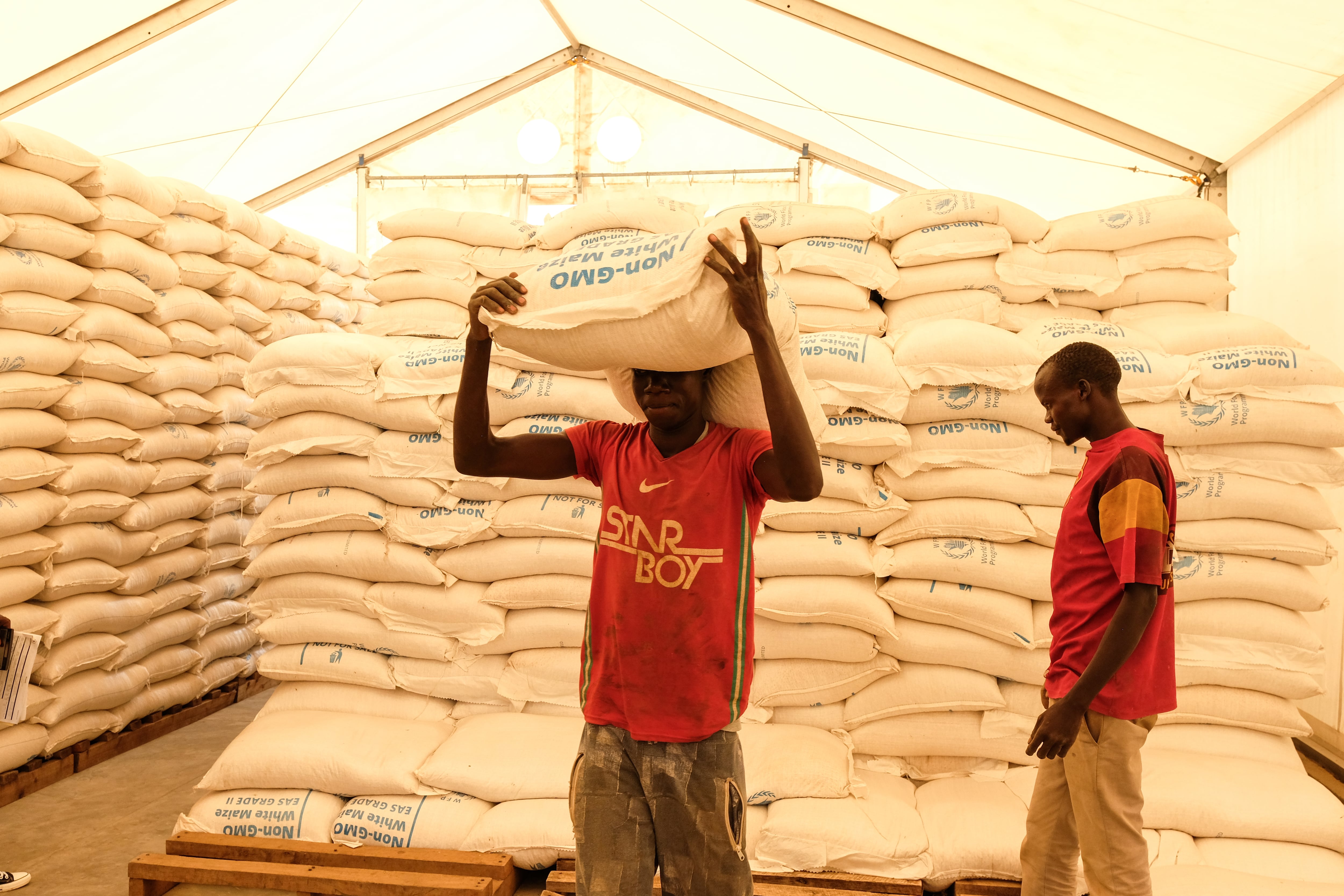 Two men stand next to stacks of relief food.