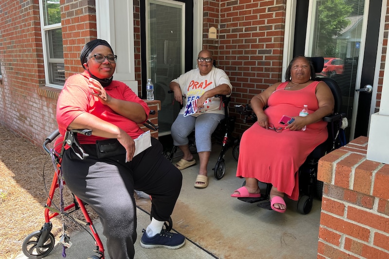 Three women sit in chairs outside a red brick building.