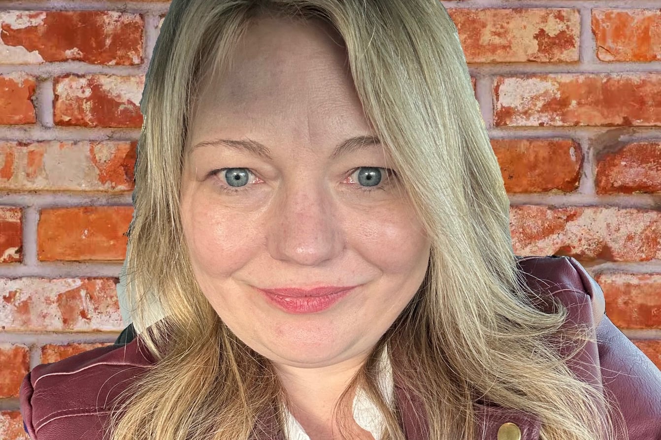 A woman with long blonde hair smiles for a portrait in front of a red brick wall.