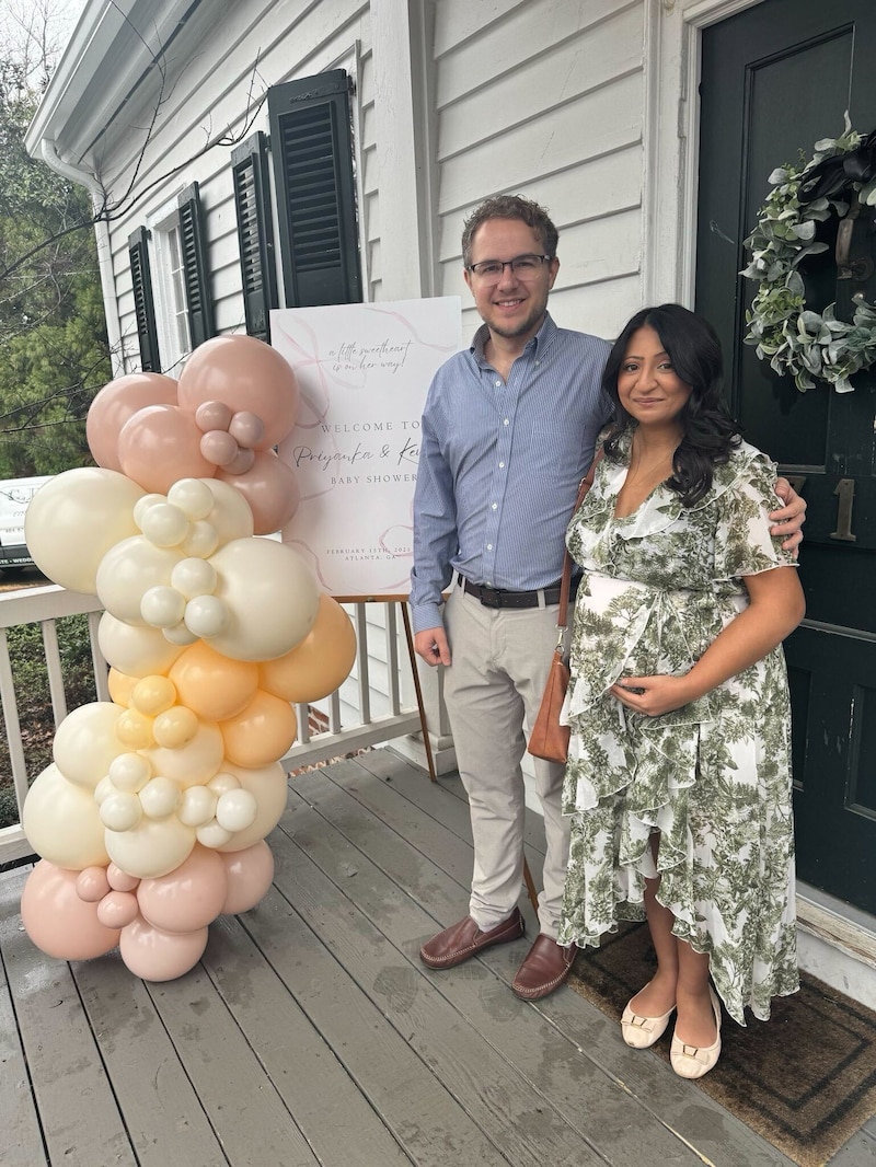A couple stand on a porch of a home next to balloons.