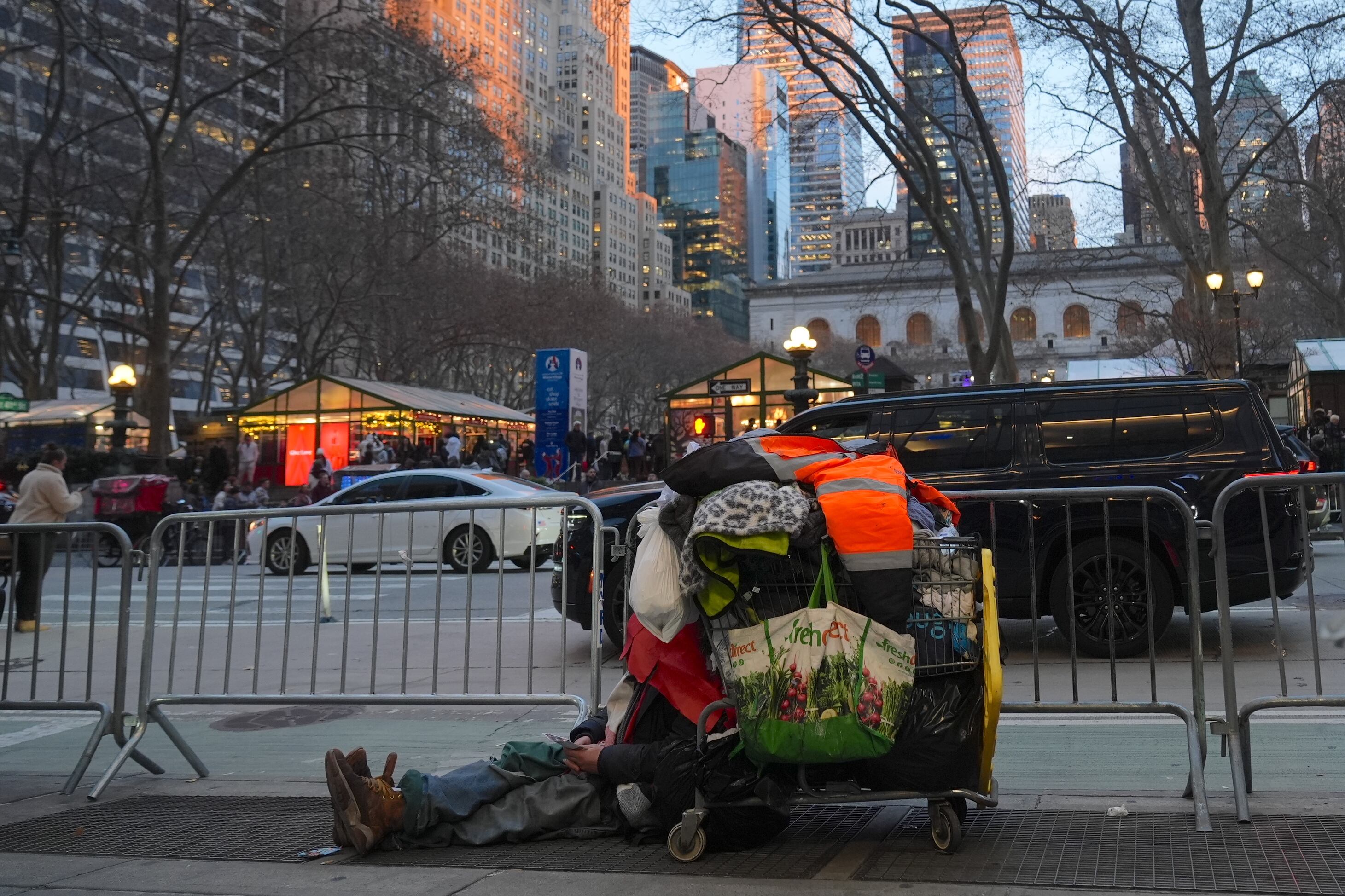 A homeless person rests on the ground next to an overloaded cart.
