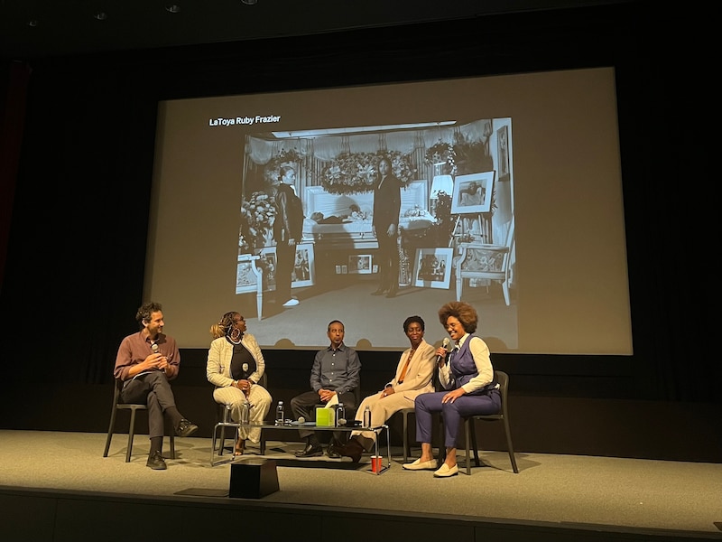 A panel made up of an artist and four public health experts hold a discussion on a state with a photo on a large screen in the background.