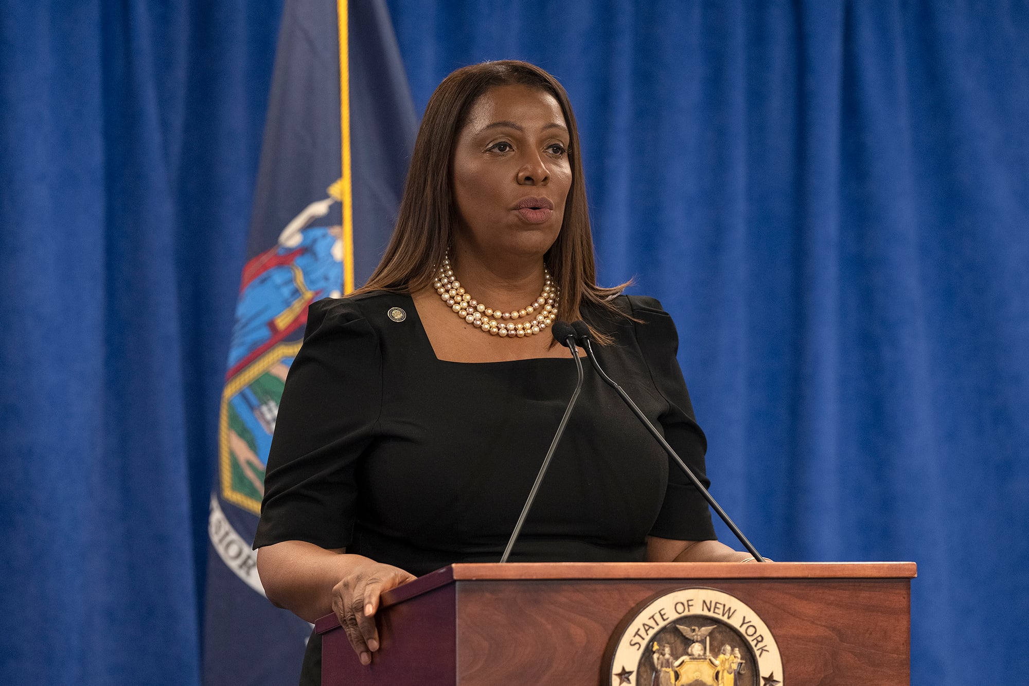 A Black woman stands behind a podium and in front of a large blue background and a New York State flag.