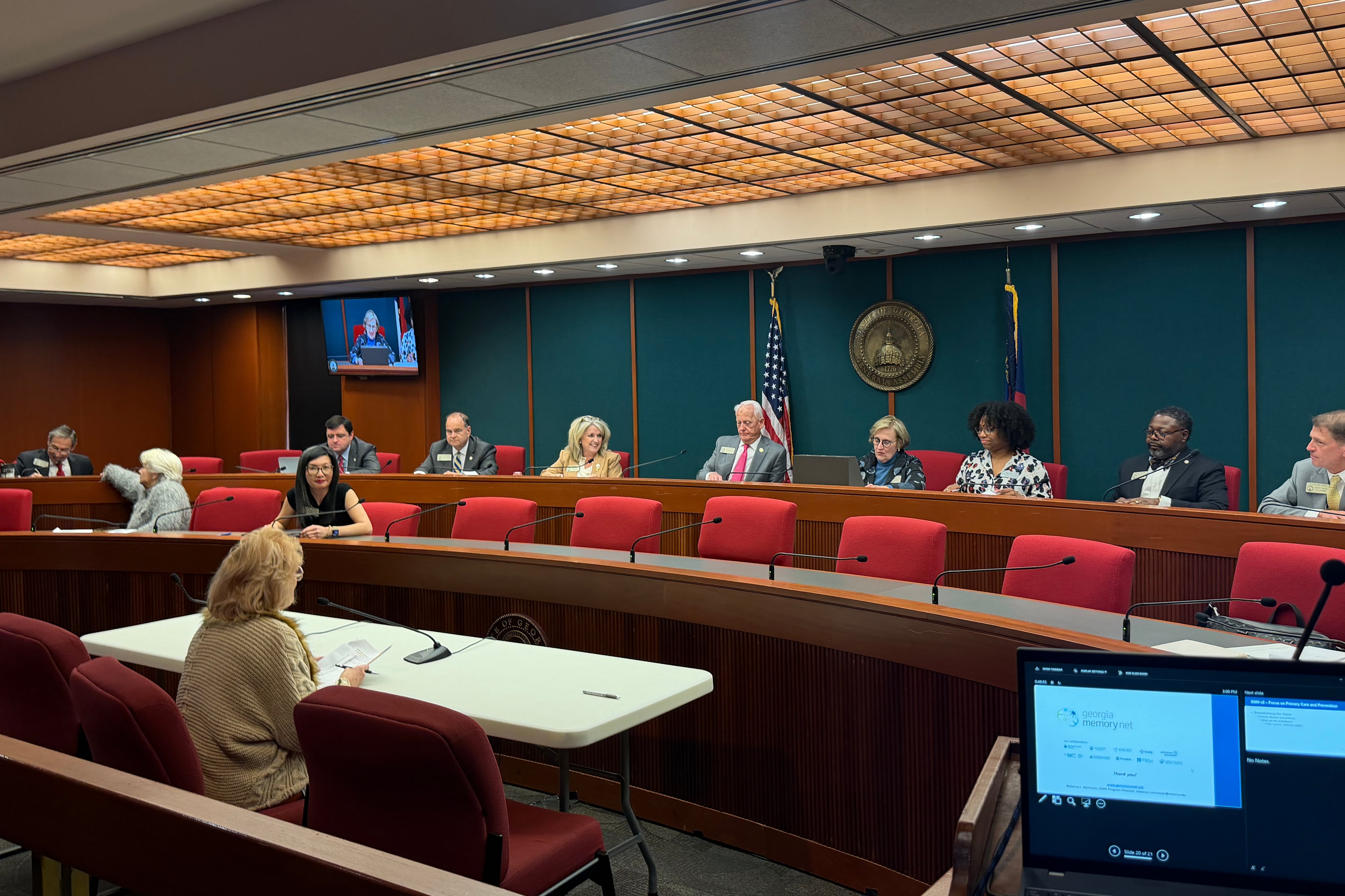 A group of legislators in a committee room face a woman in tan clothing sitting at a table with a microphone.