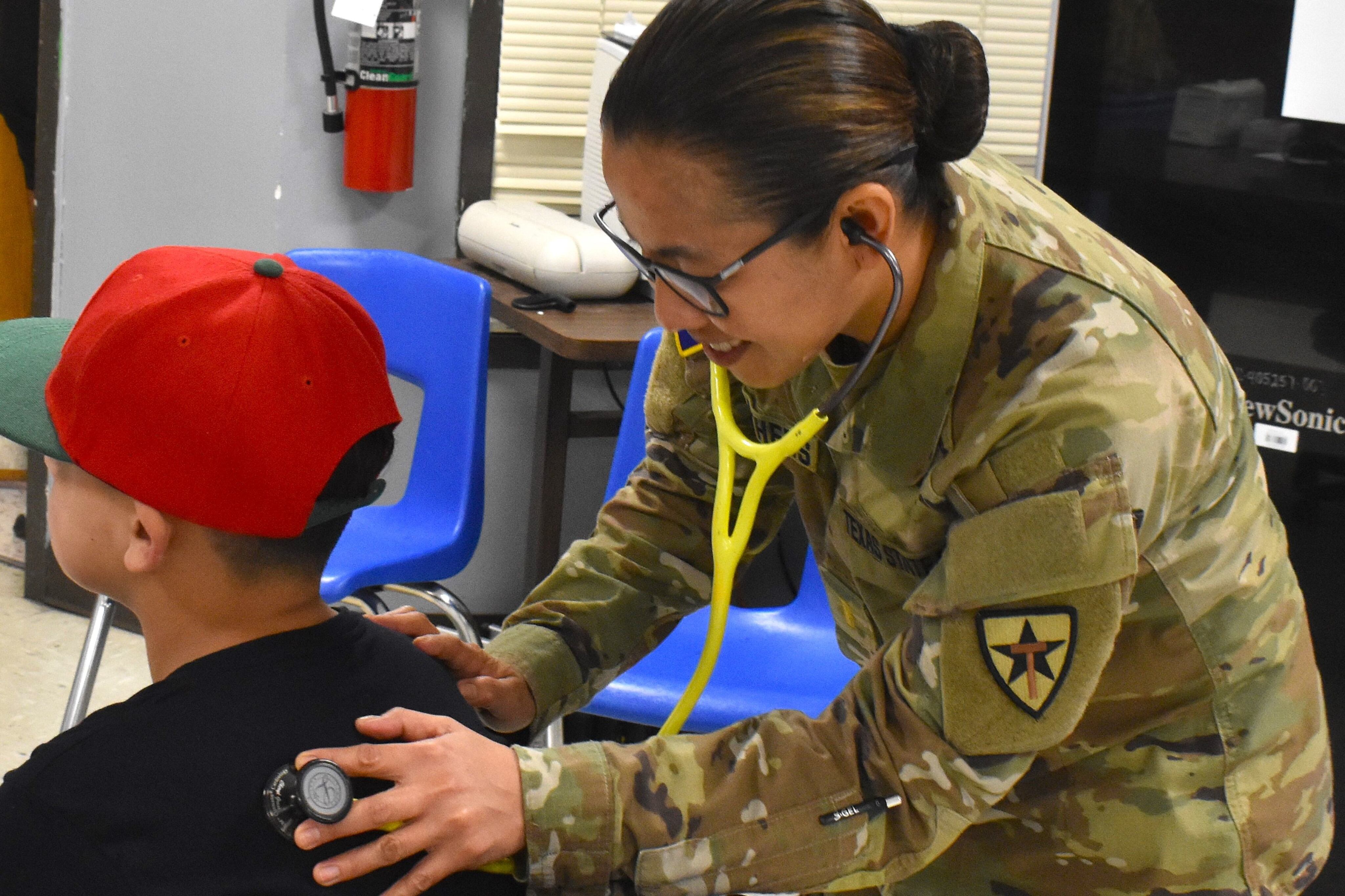 A woman wearing a camouflage Texas State Guard uniform listens to a man's back with a stethoscope.