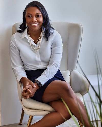 A Black woman with short dark hair and wearing a white blouse poses for a portrait while sitting on a white chair.