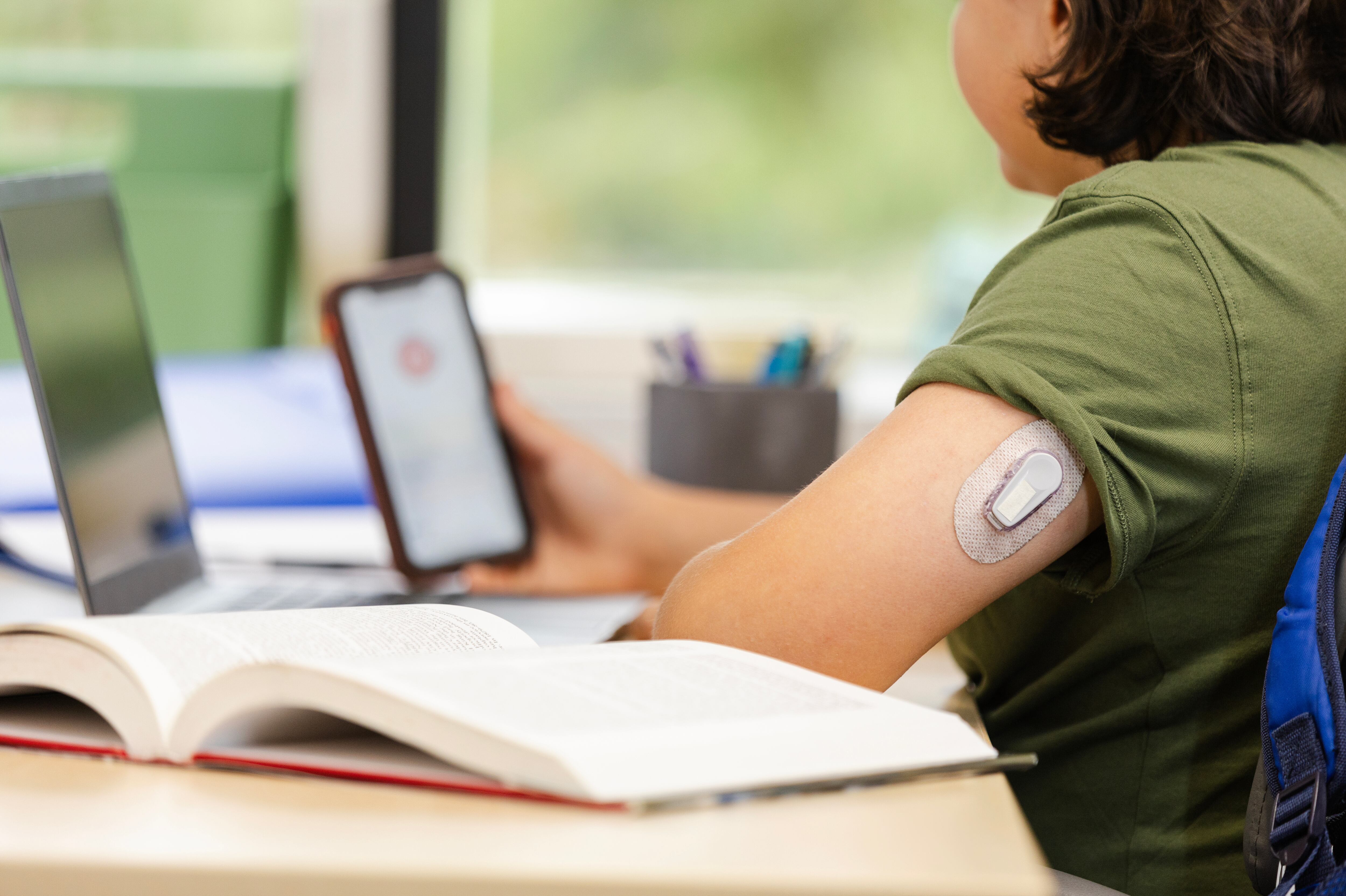 A child looks at a glucose monitoring app on a cellphone near an open laptop and book.