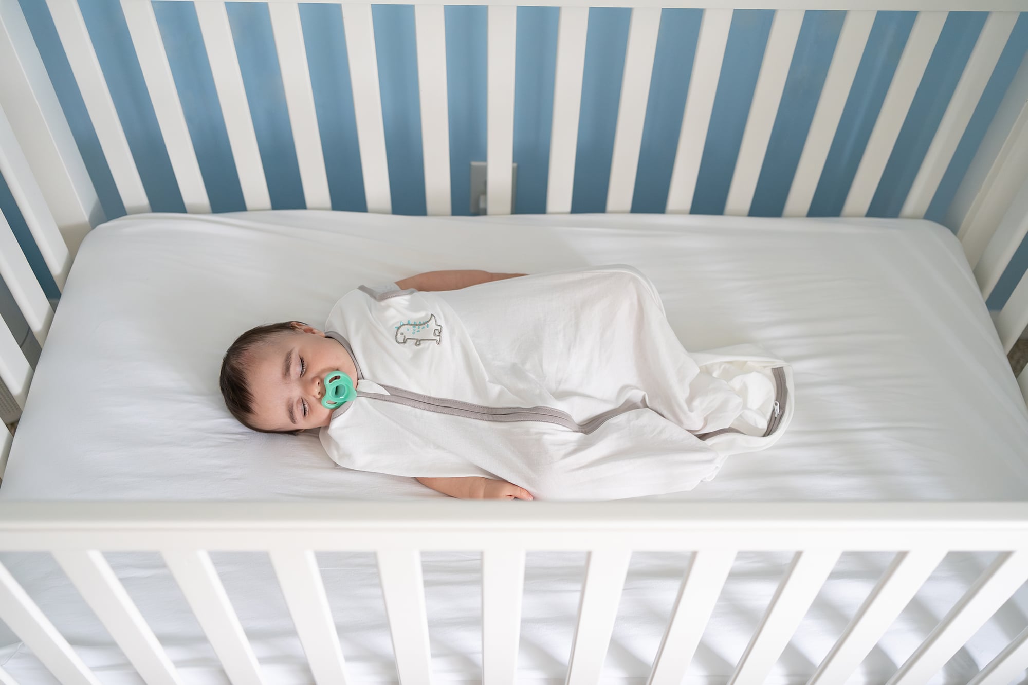 A small baby in a white sleeper onesie and with a blue pacifier in their mouth sleeps on their back in a white cribe.