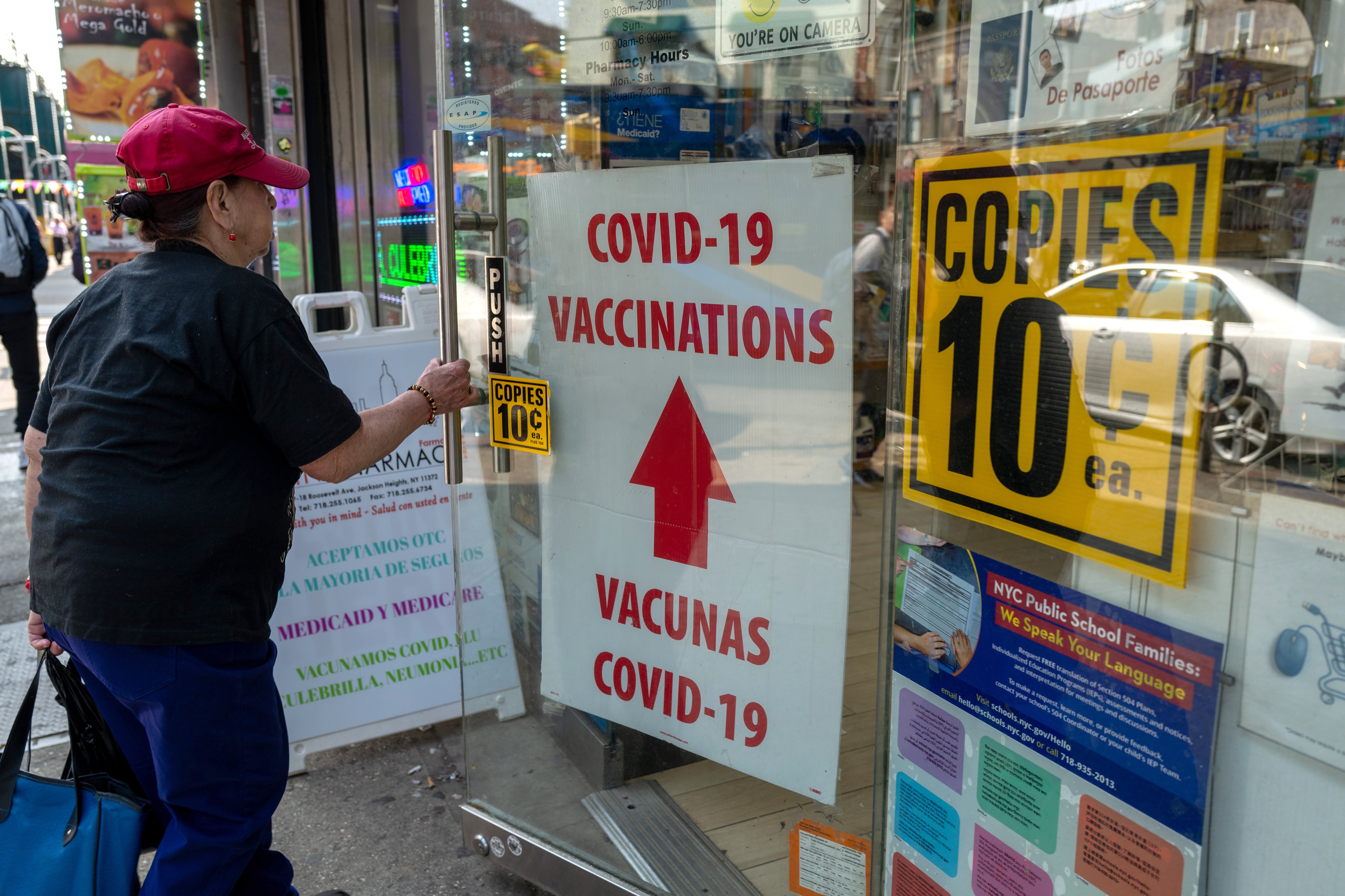 A person opens a glass door to a pharmacy with posters and signs attached to the side of the glass door and windows.