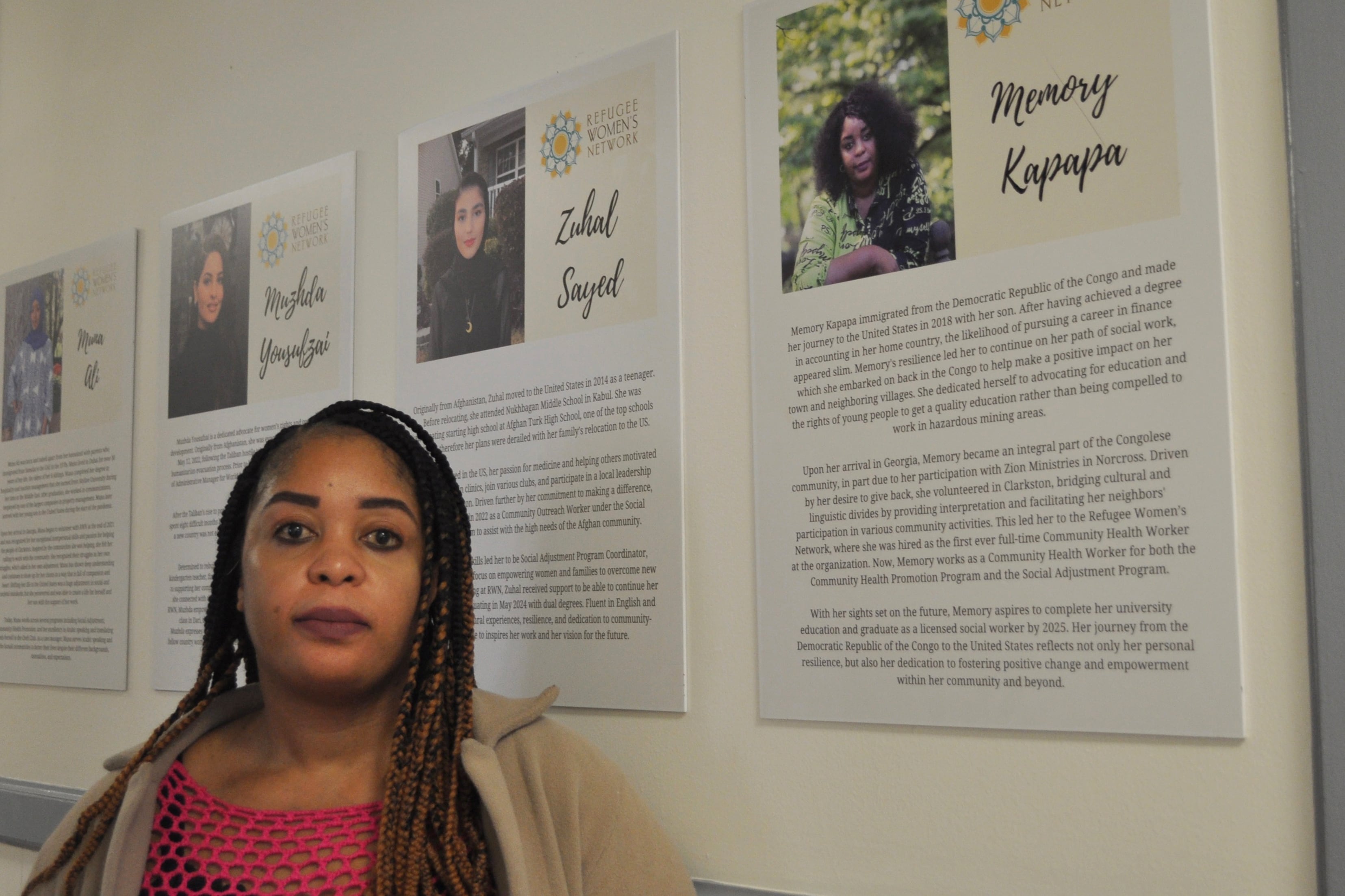 A Black woman with long, dark hair poses for a portrait next to a wall full of info posters.
