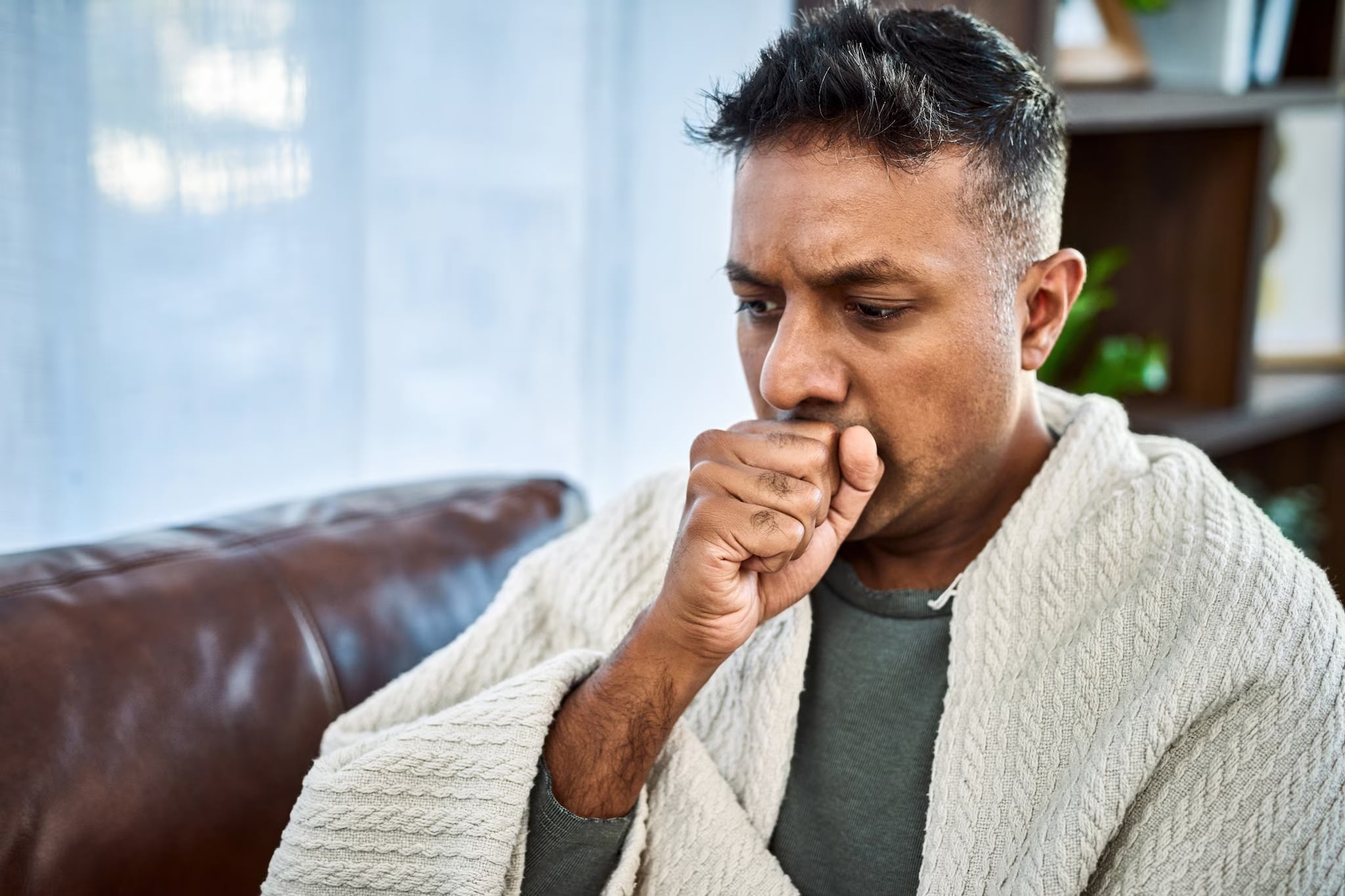 A man covered by a blanket sits on a couch and coughs into his fist.