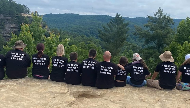 A group of people sit along a cliff outdoors with their backs to the camera, wearing black T-shirts that read "This is what recovery looks like."