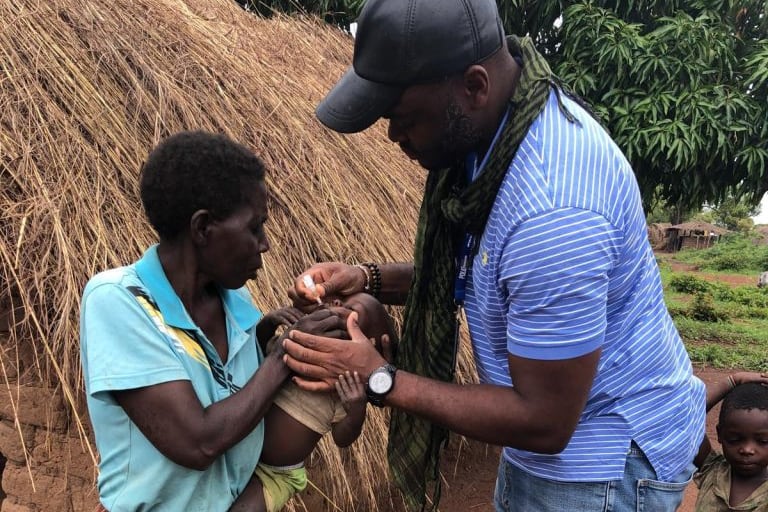 A man vaccines a baby held by another person next to a hut.