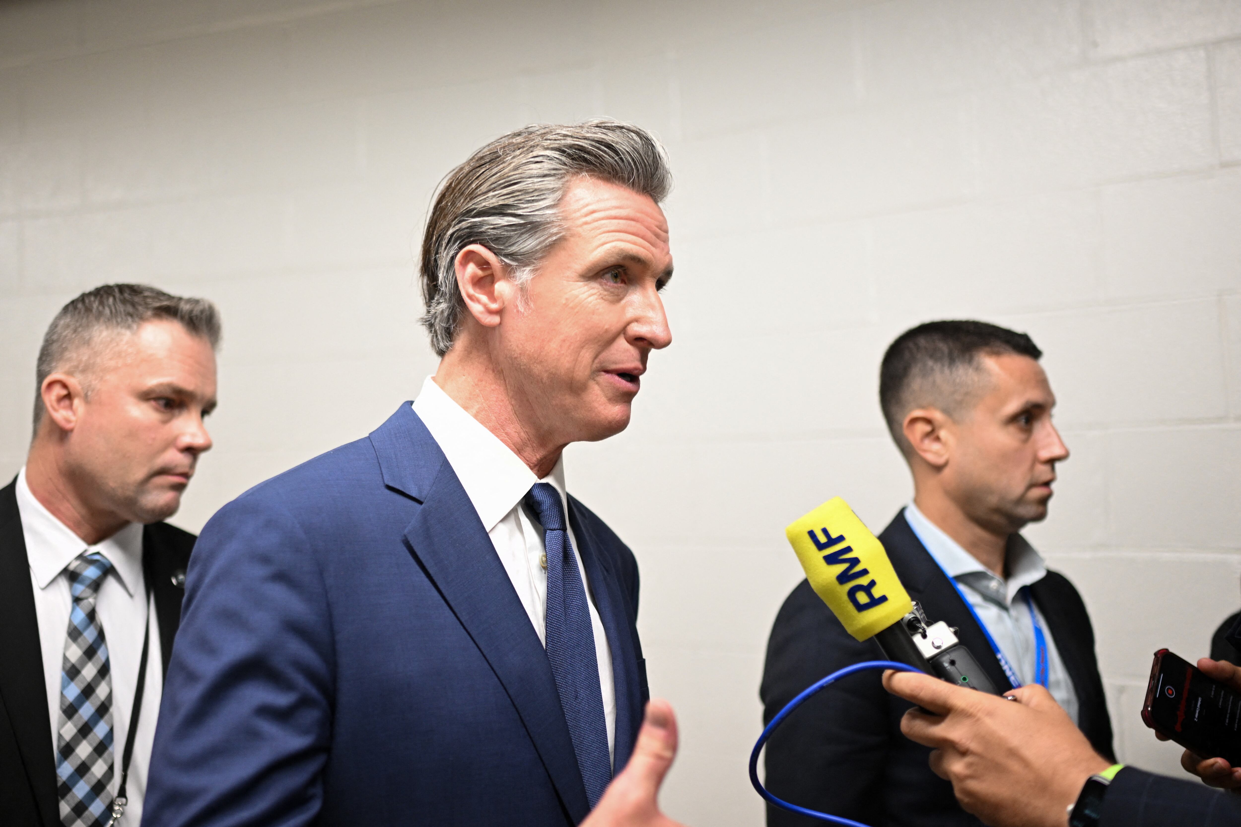 California Governor Gavin Newsom, surrounded by two other men, speaks into a microphone in a hallway.