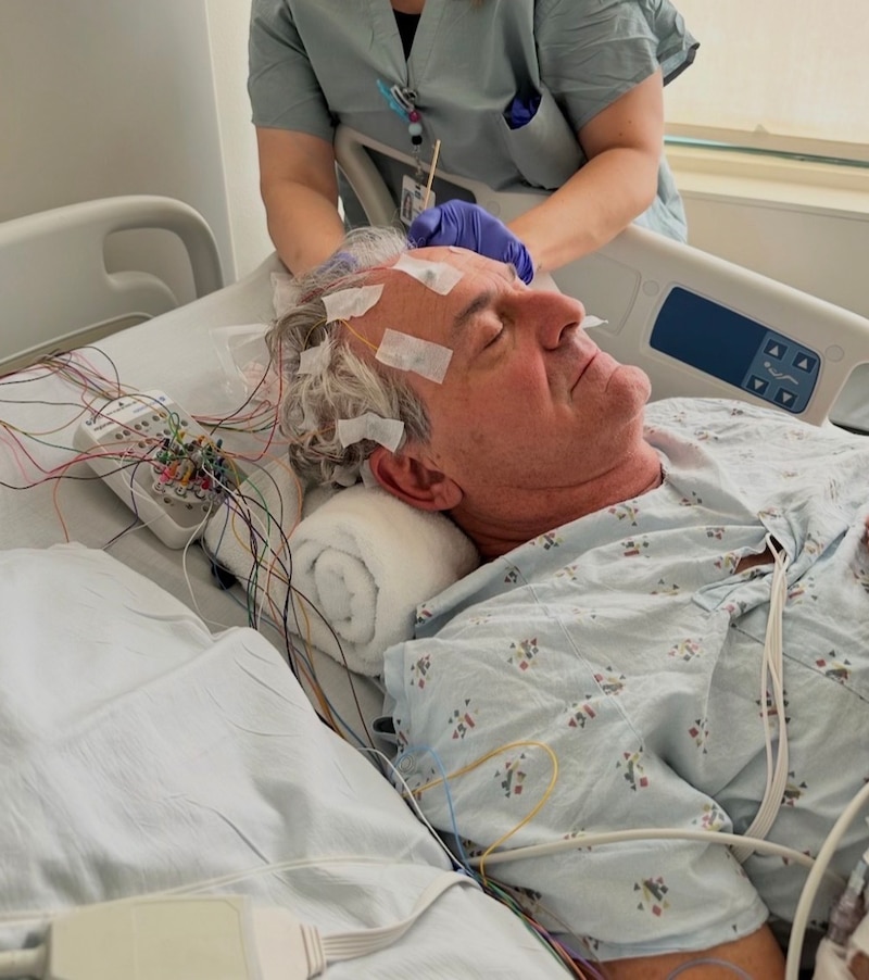 A man in a hospital gown with electrodes on his head is lying down on a bed while a woman adjusts the electrodes.