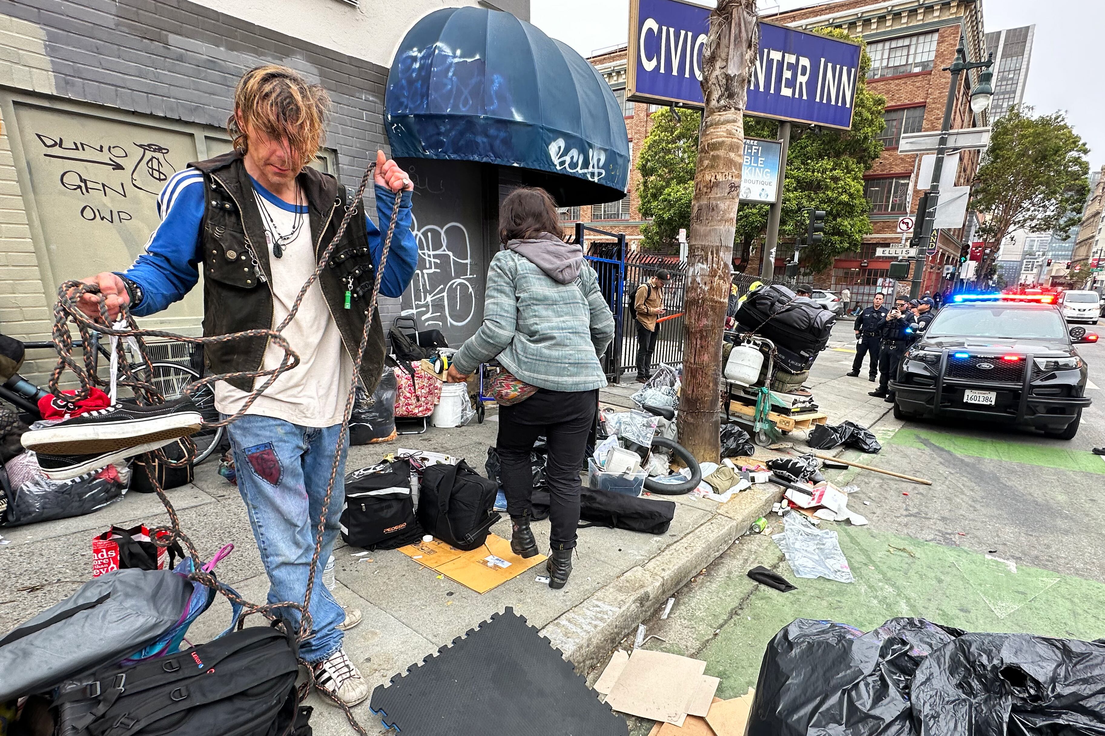 Homeless people gather their belongings on the street, with a police car in the background.