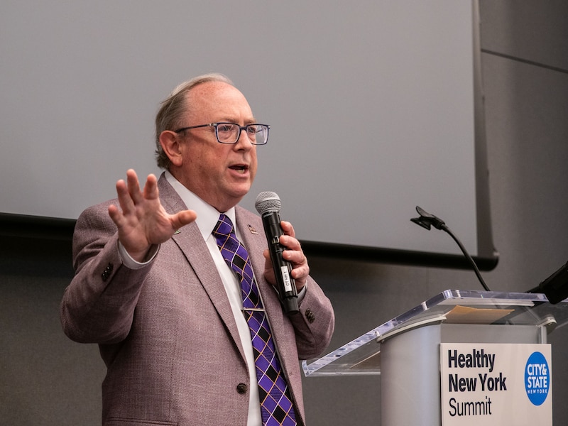 A white man with glasses and wearing a tan suit speaks from a podium in a conference room.