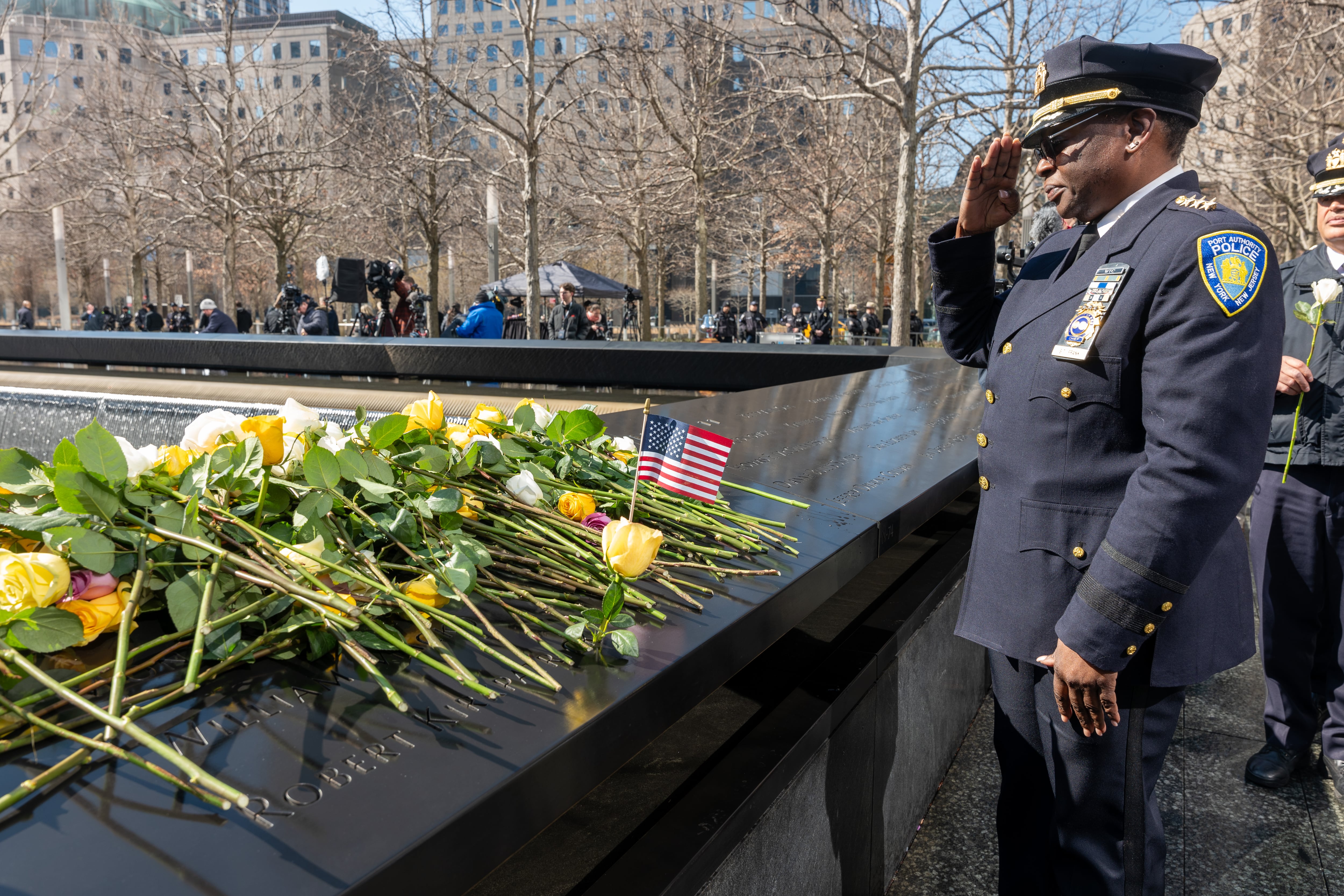A police officer in uniform salutes at the 9/11 memorial covered in flowers.