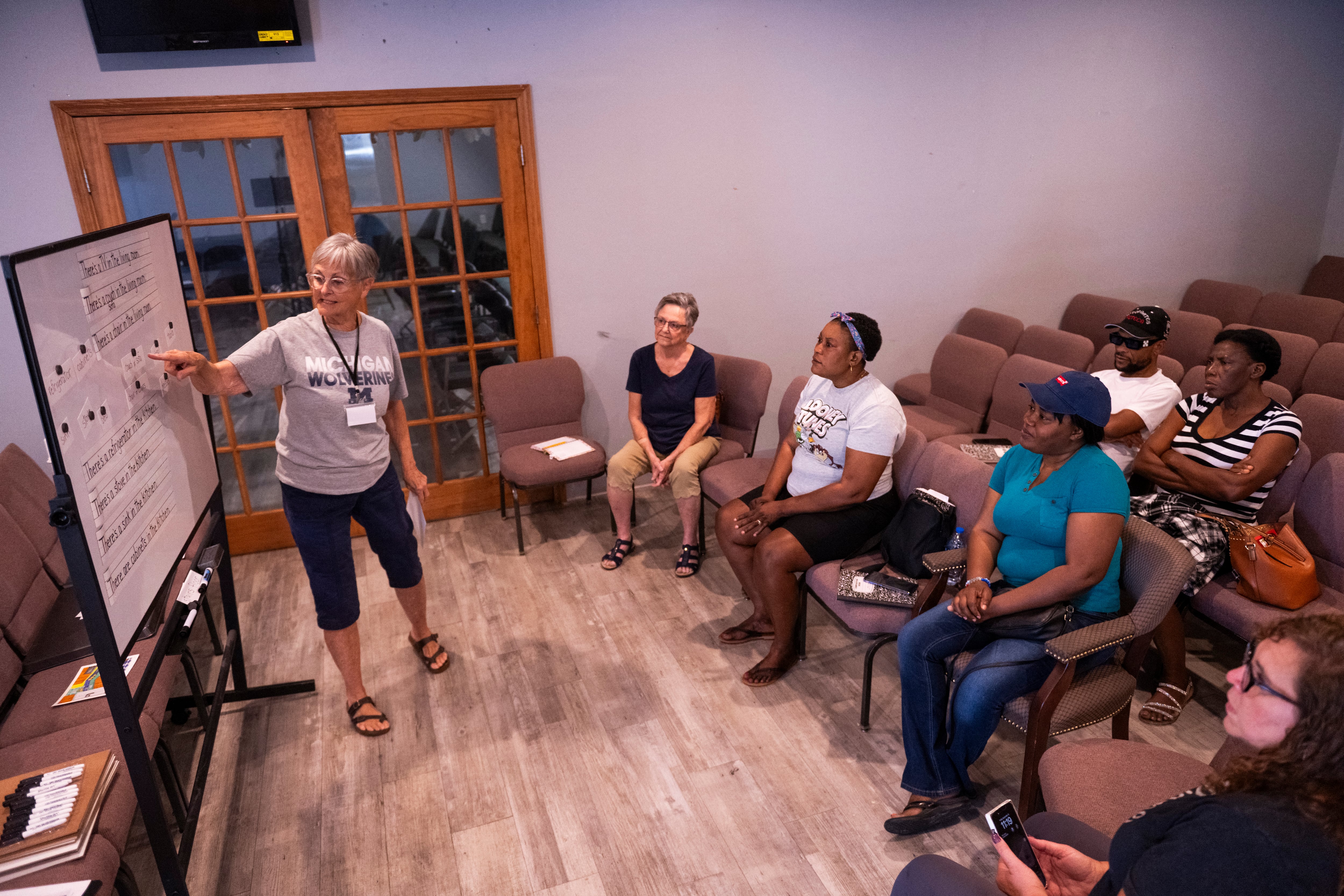 A volunteer stands and points to a whiteboard in front of a group of immigrants seated in a classroom setting.