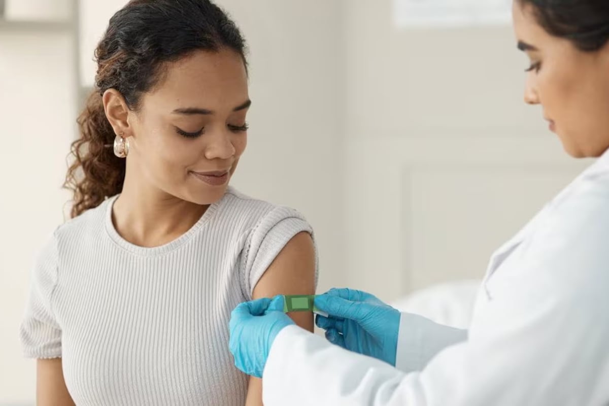 A doctor places a band-aid on the arm of a woman with a rolled-up short-sleeve t-shirt.