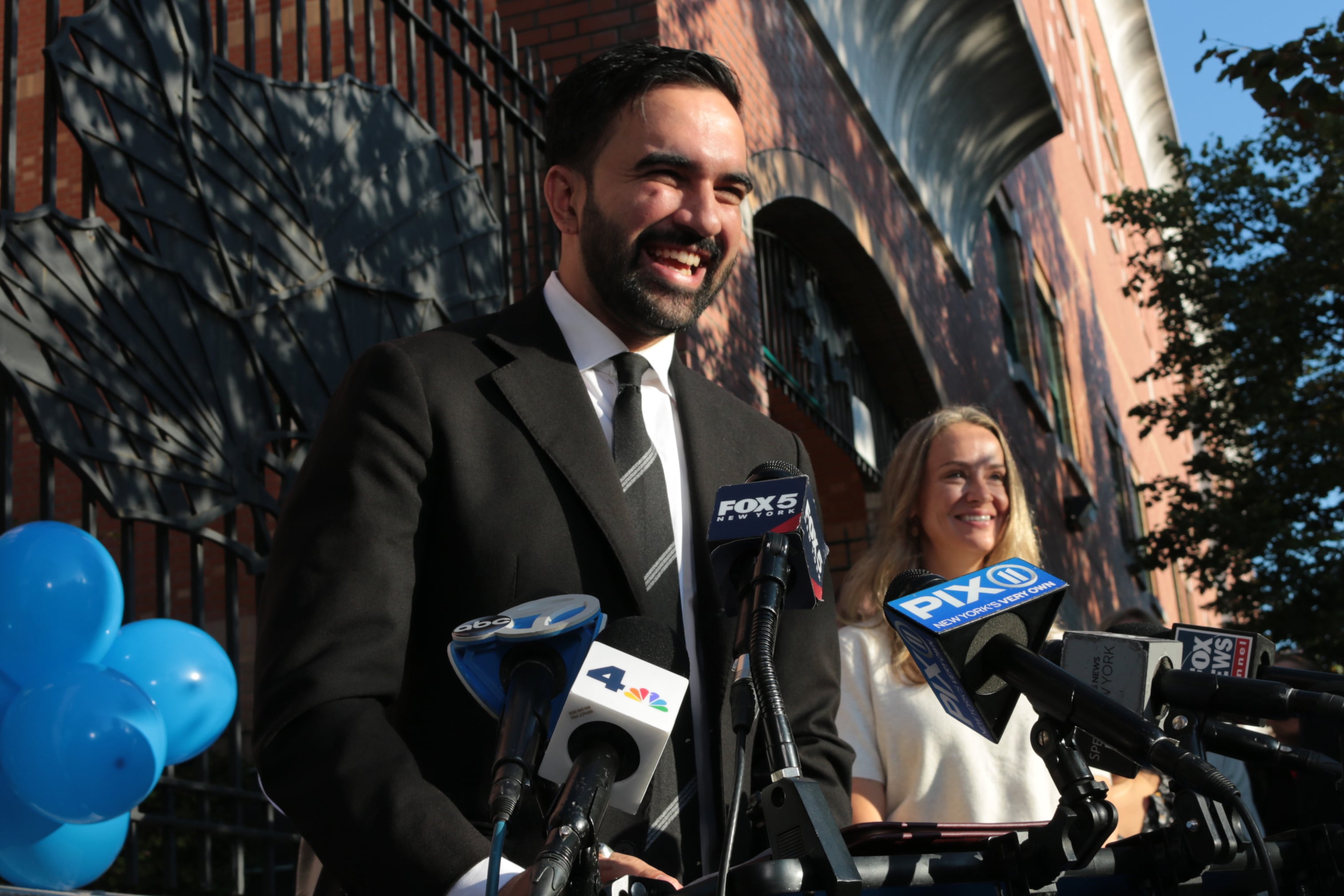 A photograph of a man in a suit standing behind a group of microphones outside.