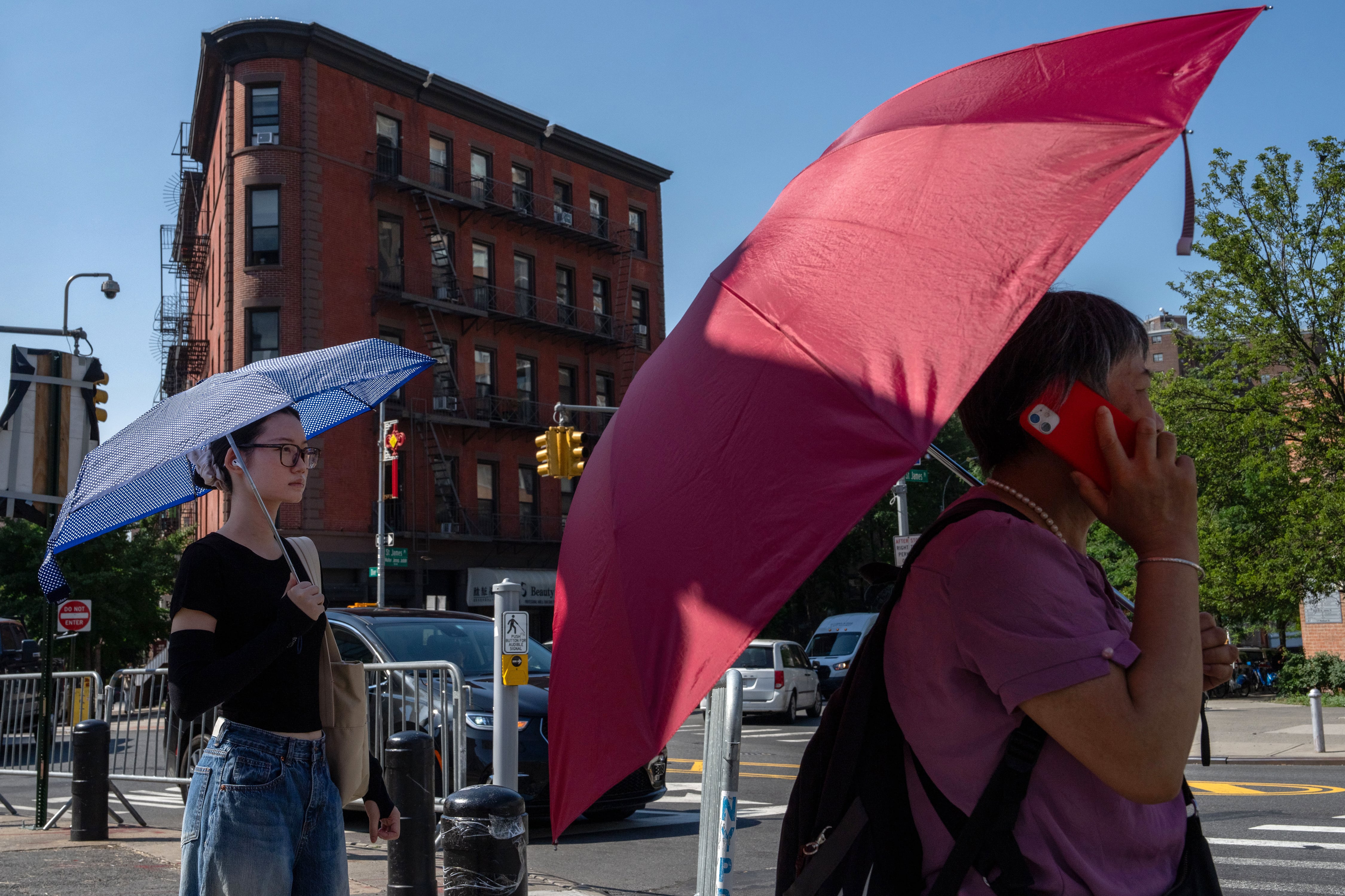 Two people holding umbrellas outside on a hot and sunny day with a large brick apartment in the background.