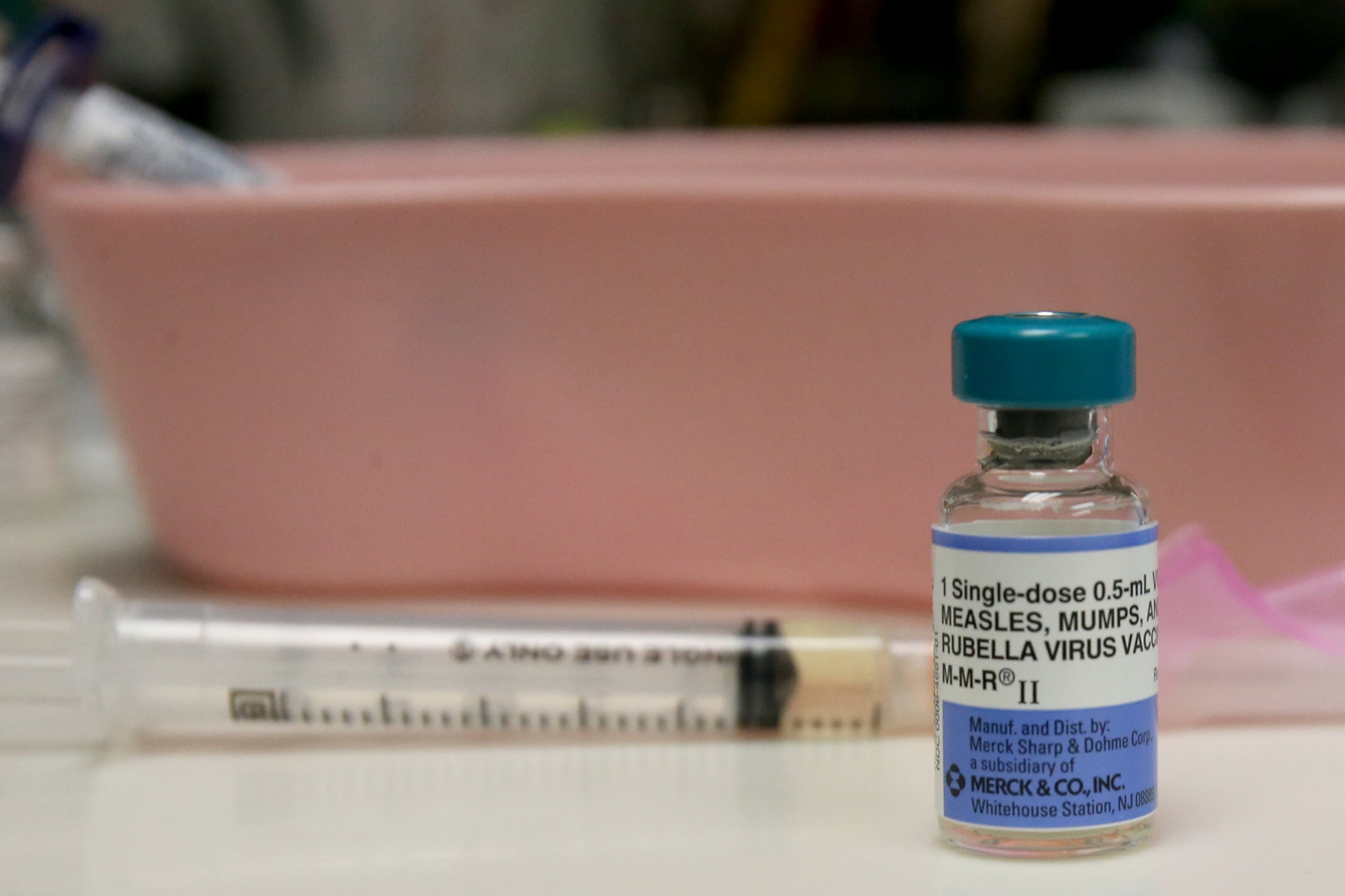 A needle and a vile of measles, mumps, rubella vaccine sit on a table in front of a light pink container.