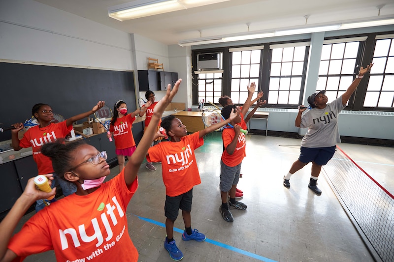 A group of children all wearing orange t-shirts stand with one of their hands in the air while an adult stands at the front of the room.