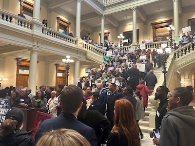A large group of people holding signs protest on the stone staircase of a large state Capitol building.