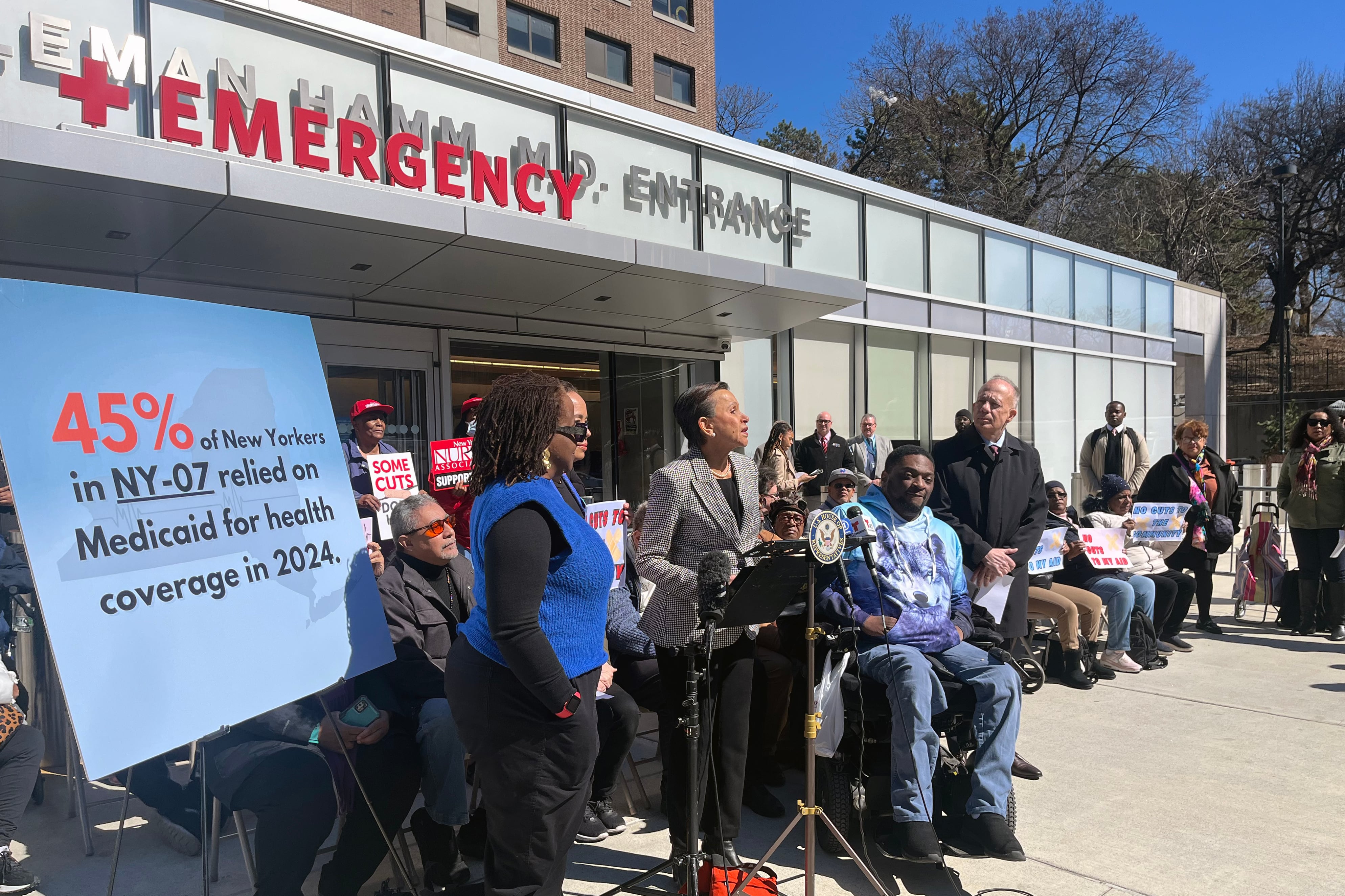 A group of people, some standing and sitting are gathering for a press conference outside the front entrance of an emergency room on a sunny day.