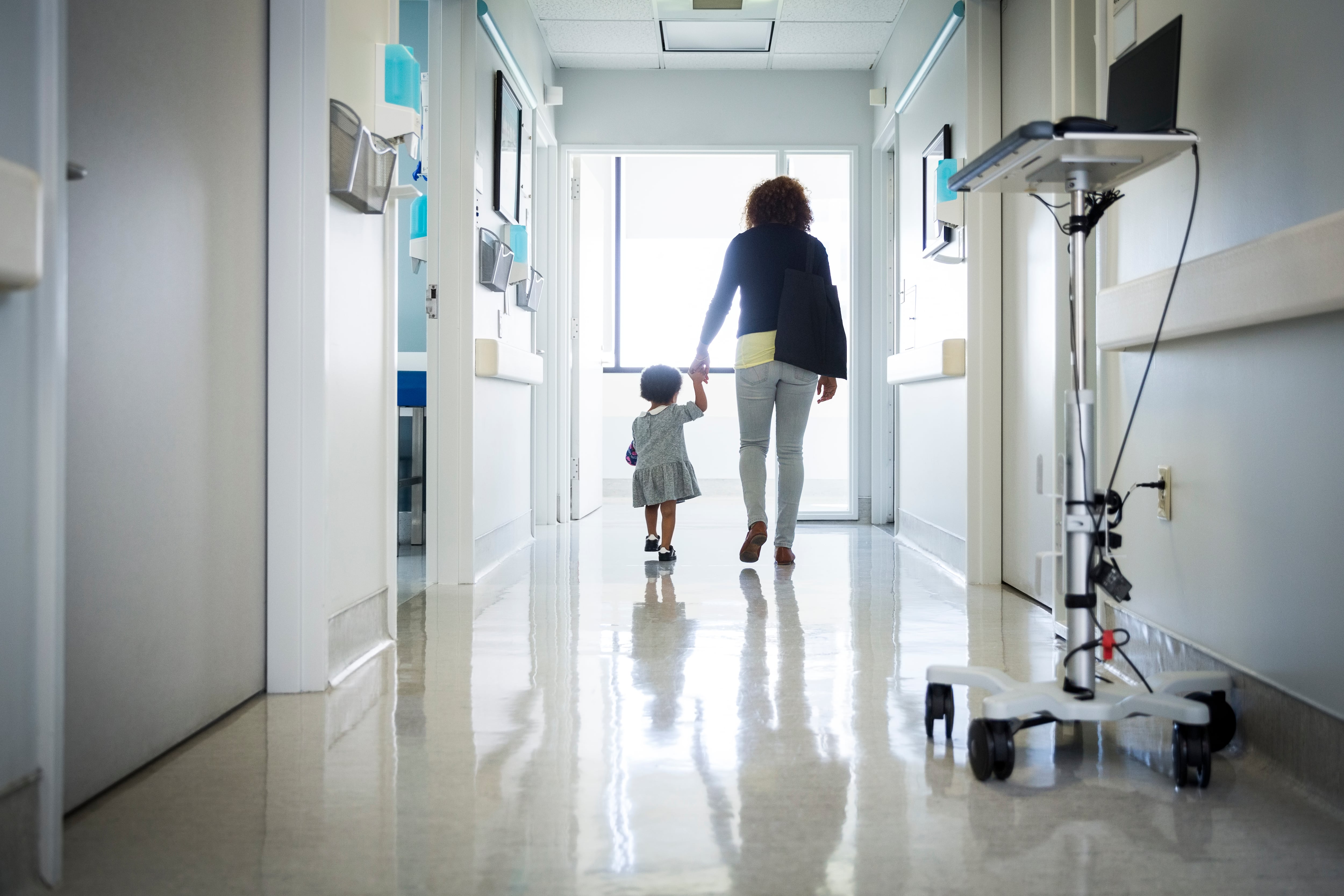 A woman holds hands with a young child as they walk along a hospital corridor.