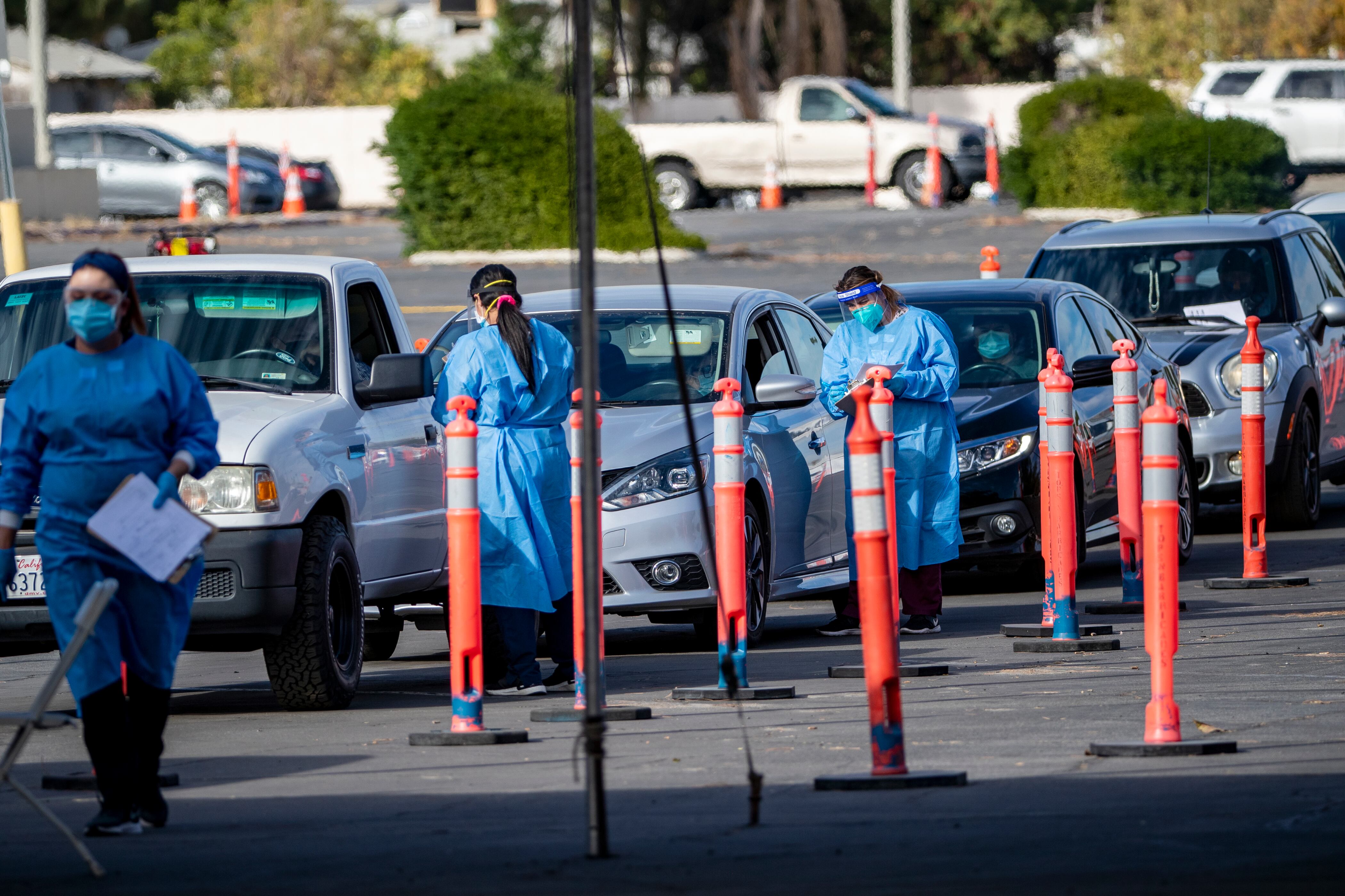 A line of cars in a parking lot with three healthcare workers dressed in blue PPE gear stand next to the car windows and tall, orange caution cones are visible on the side.