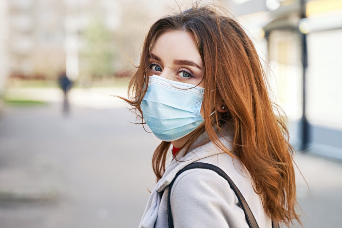A woman wearing a hospital mask over her face stands outside with a purse strapped around her shoulder.
