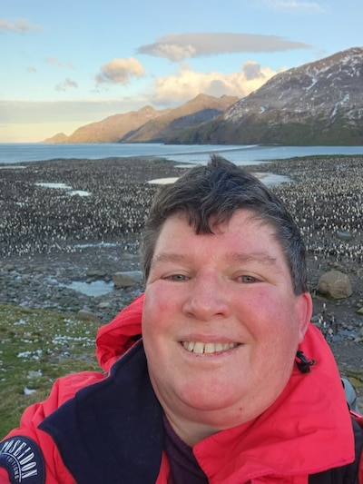 A woman with short dark hair wearing a red parka takes a selfie with a beautiful nature scene behind her.