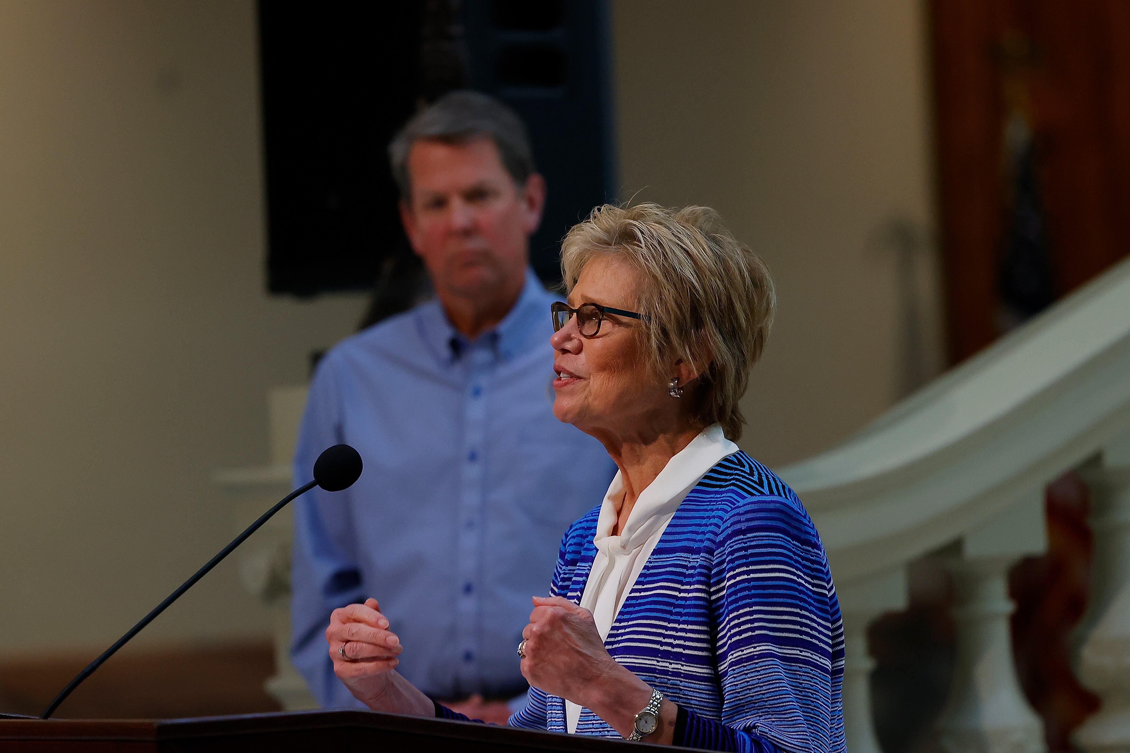 Georgia Public Health Commissioner Kathleen Toomey speaks at a podium with Gov. Brian Kemp in the background.