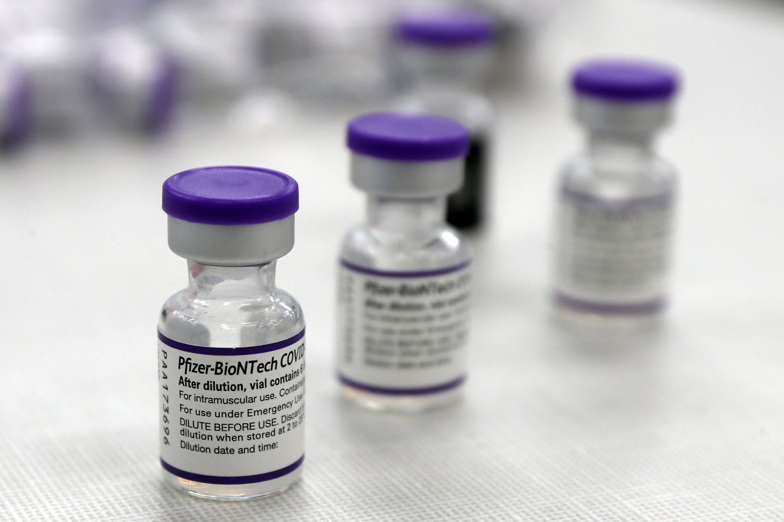 Three clear bottles with labels and purple lids sit on a table with some other bottles in the background.