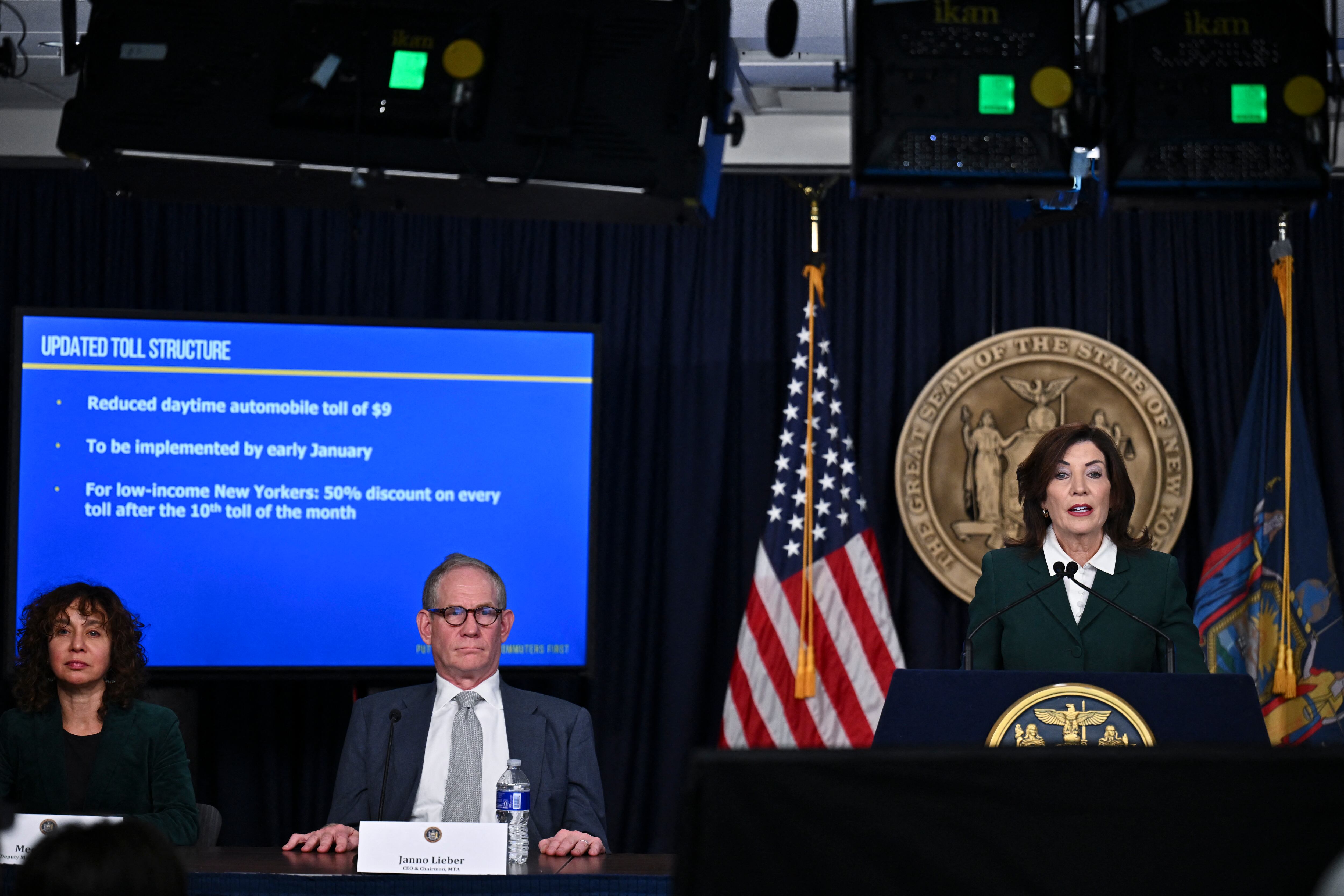 New York Governor Kathy Hochul stands at a podium with two others seated to her right.