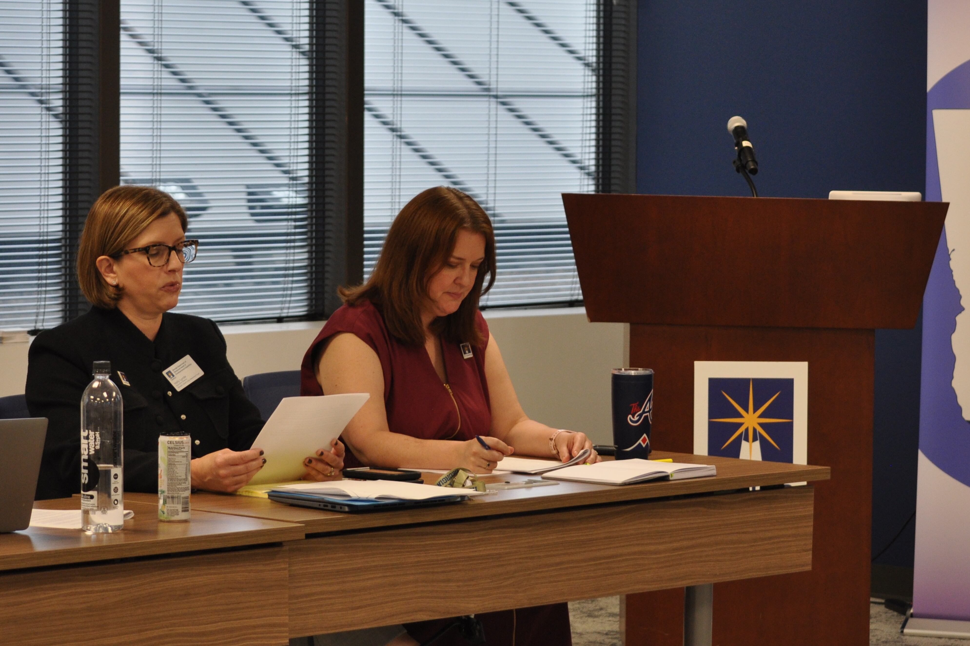 Two seated women read from papers at a table during a meeting.