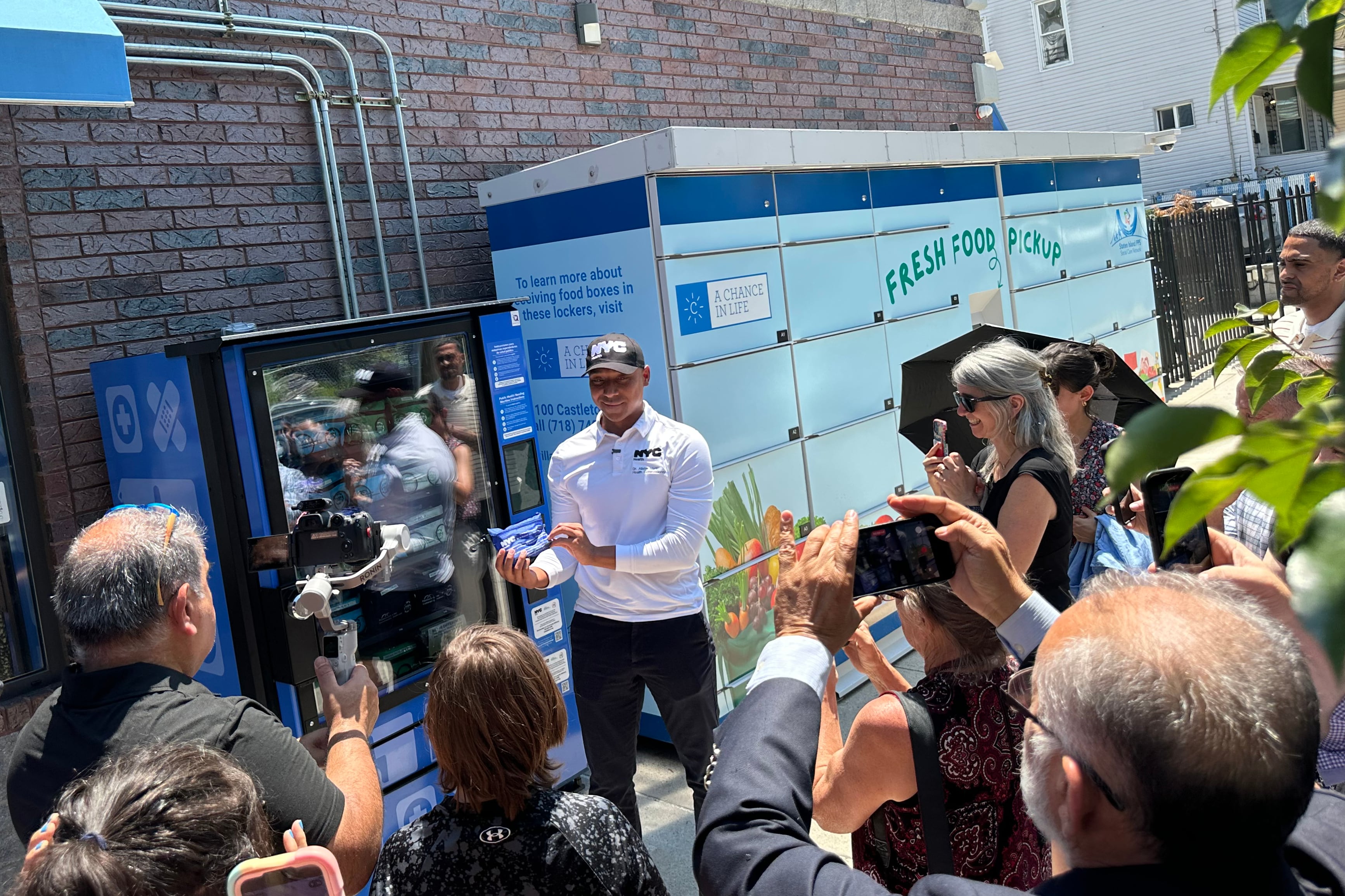 A photograph of a Black man holding a pouch outside of a vending machine while a group of people stand around him in a press conference.