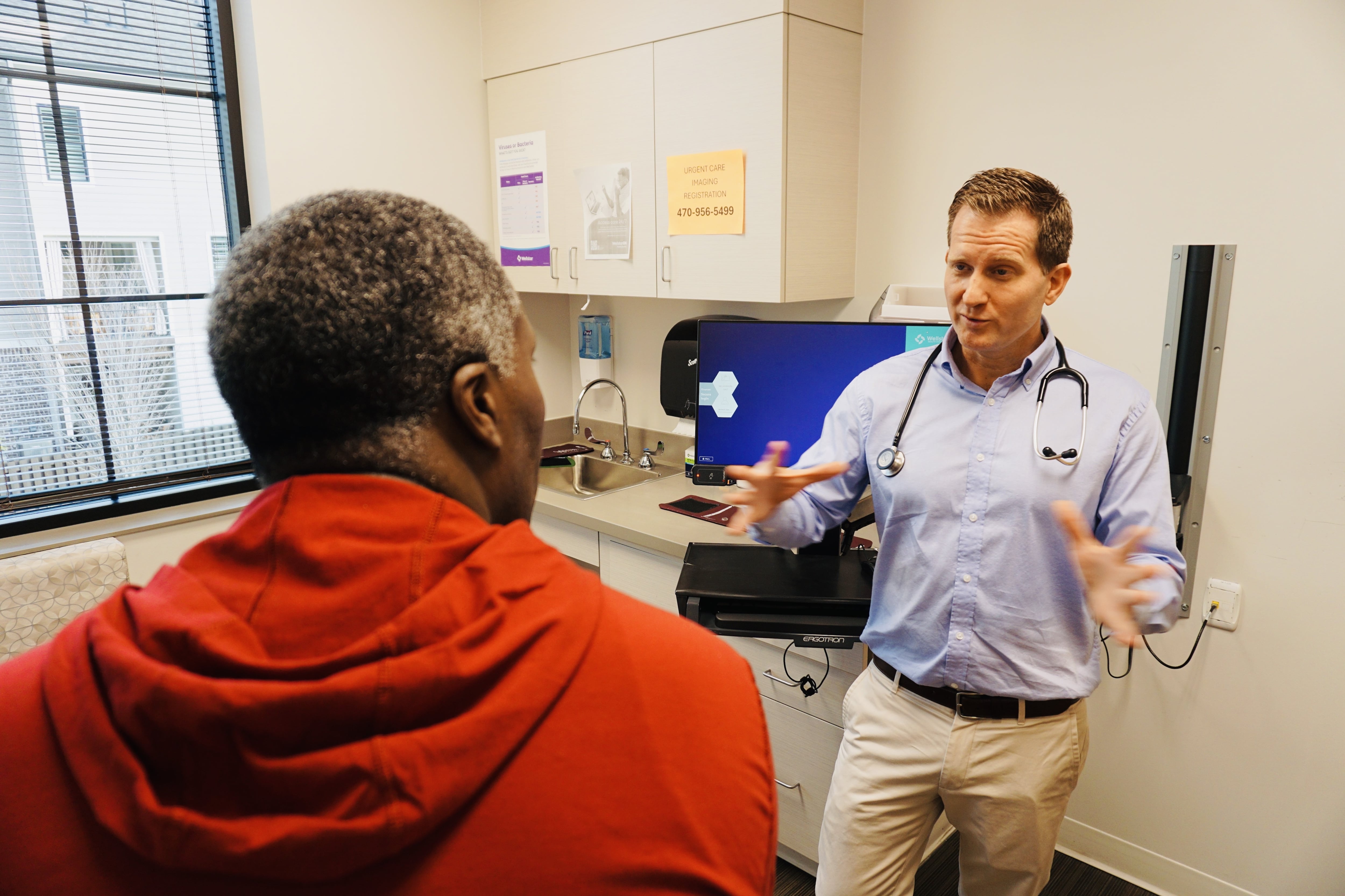 A man wearing a blue shirt with a stethoscope speaks to a patient wearing a red hooded sweatshirt a clinical exam room.