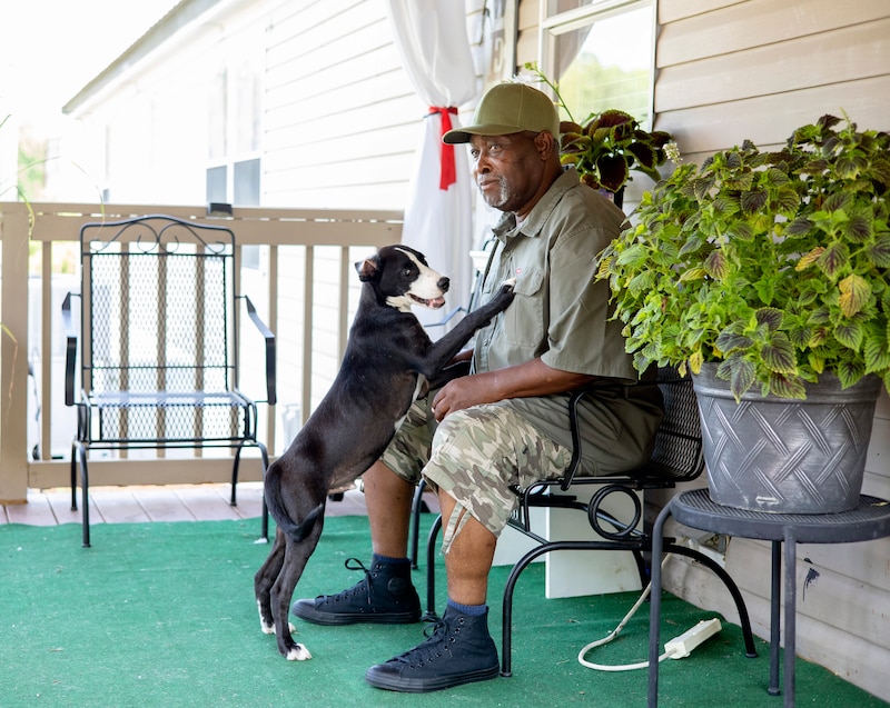 A dog stands on its hind legs to touch Joshua McCray, who is sitting on a porch.
