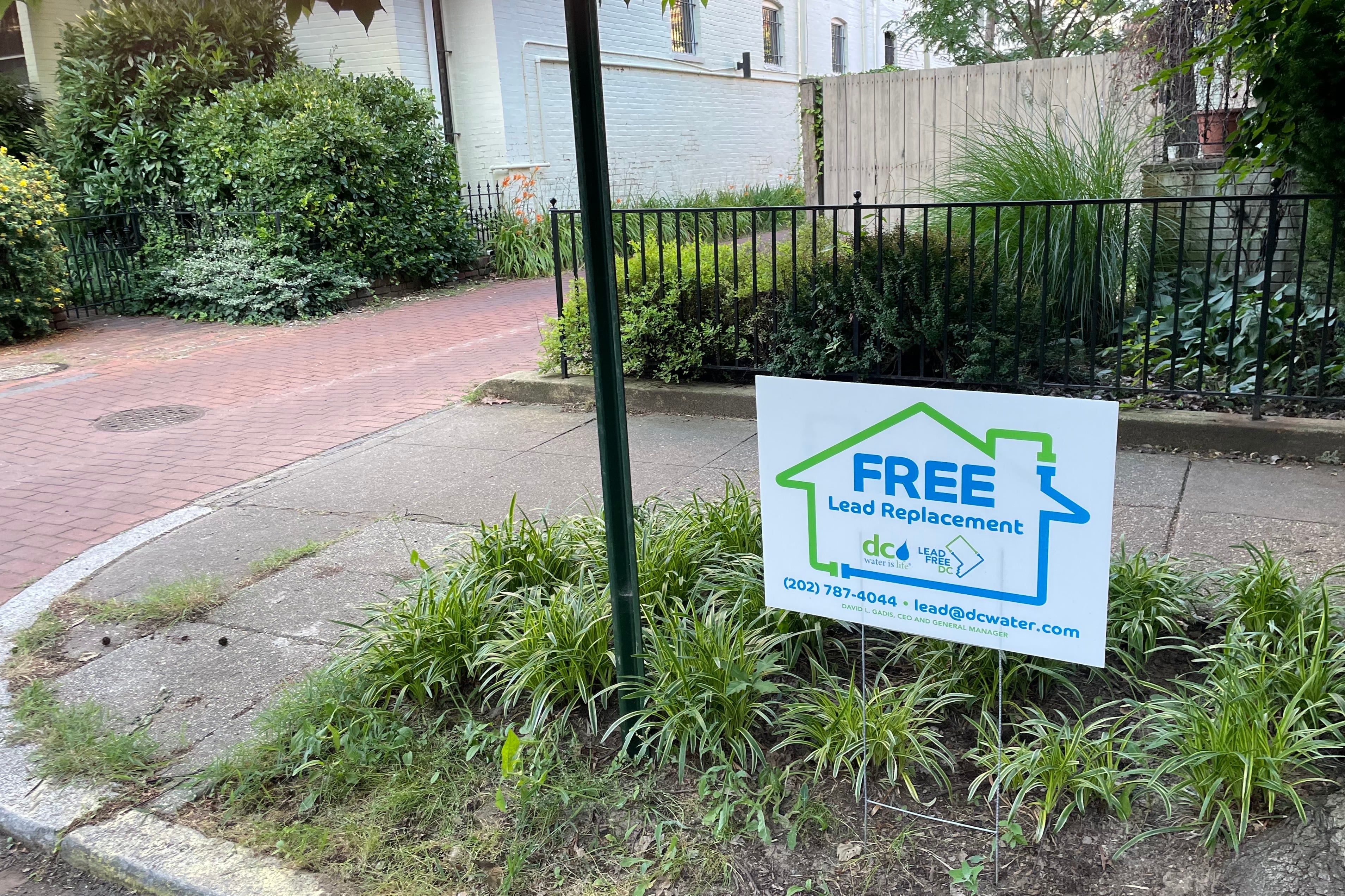 A white and blue sign sticks out of the ground near a sidewalk in front of a black fence.