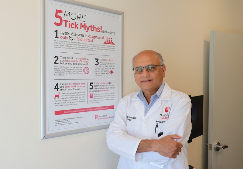 A photograph of a bald man in glasses and wearing a white doctor coat stands next to a sign on a white wall.