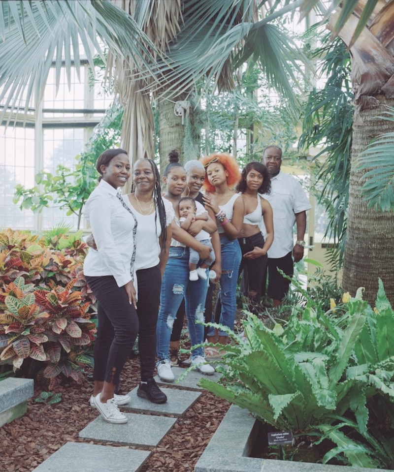Members of a family stand close together among trees and plants.